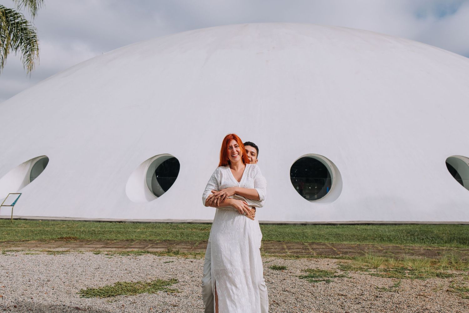 Couple in white attire posing in front of a large white dome building with circular windows outdoors.