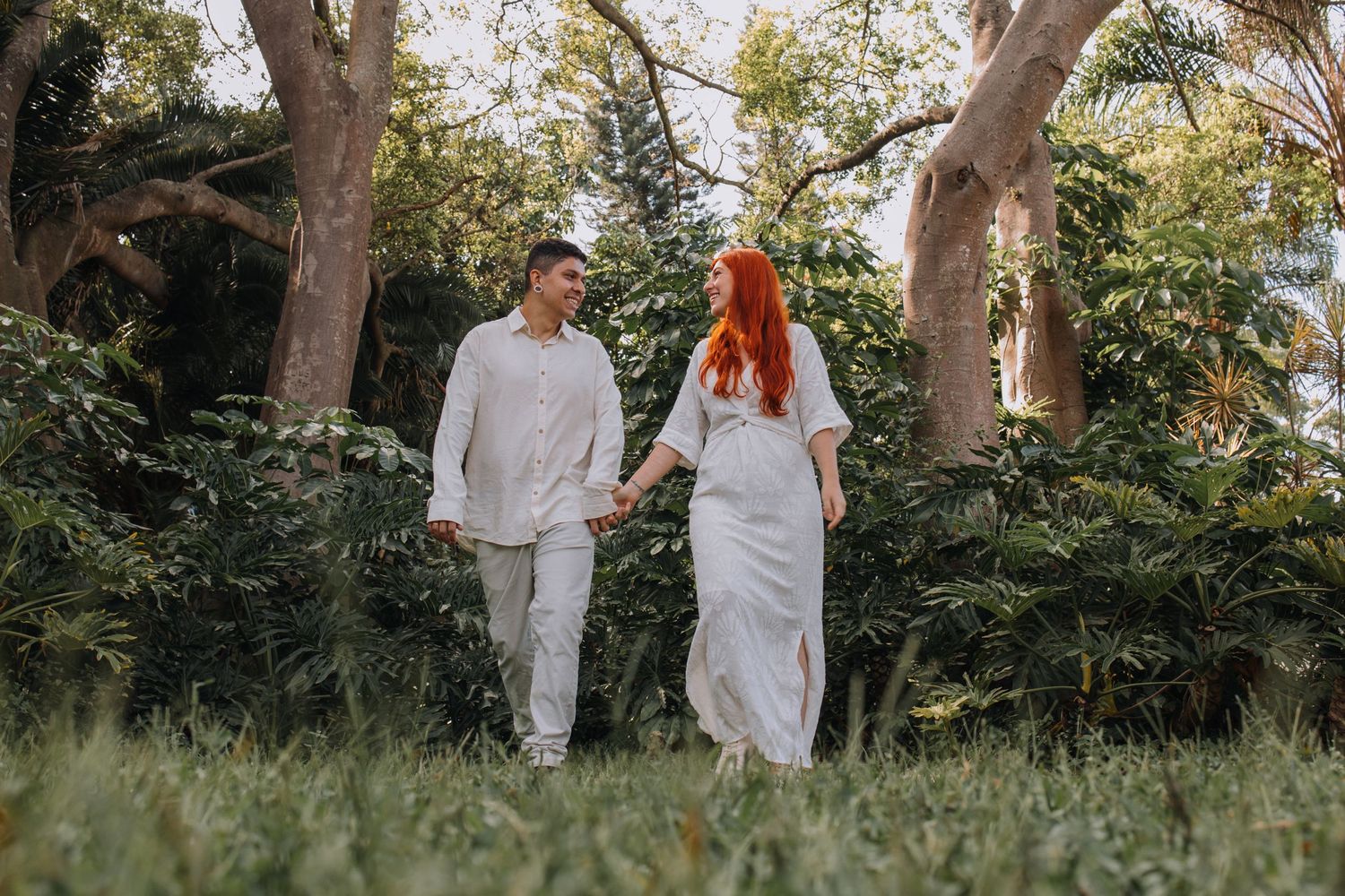 Couple in white outfits holding hands and walking through lush green park with tall trees.
