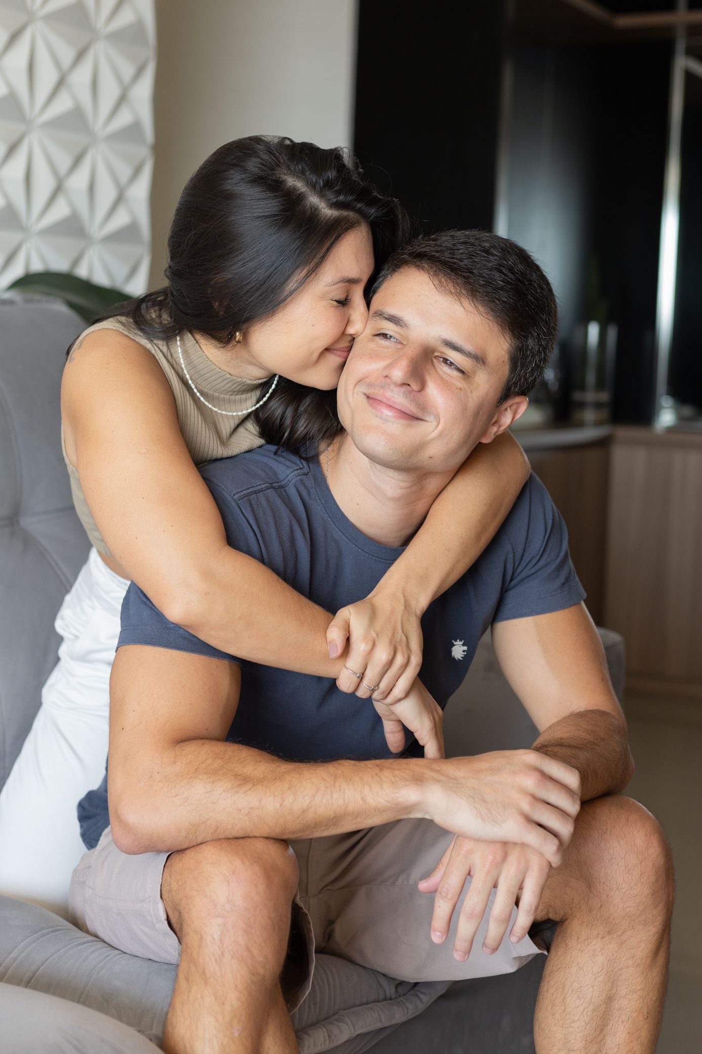 A woman hugging a smiling man from behind on a couch, kissing his cheek in a cozy living room.