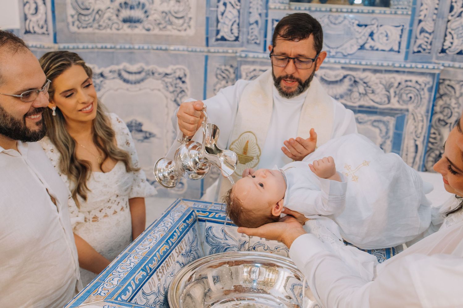 Priest baptizing a baby with holy water at a ornate blue tiled church font, surrounded by family.