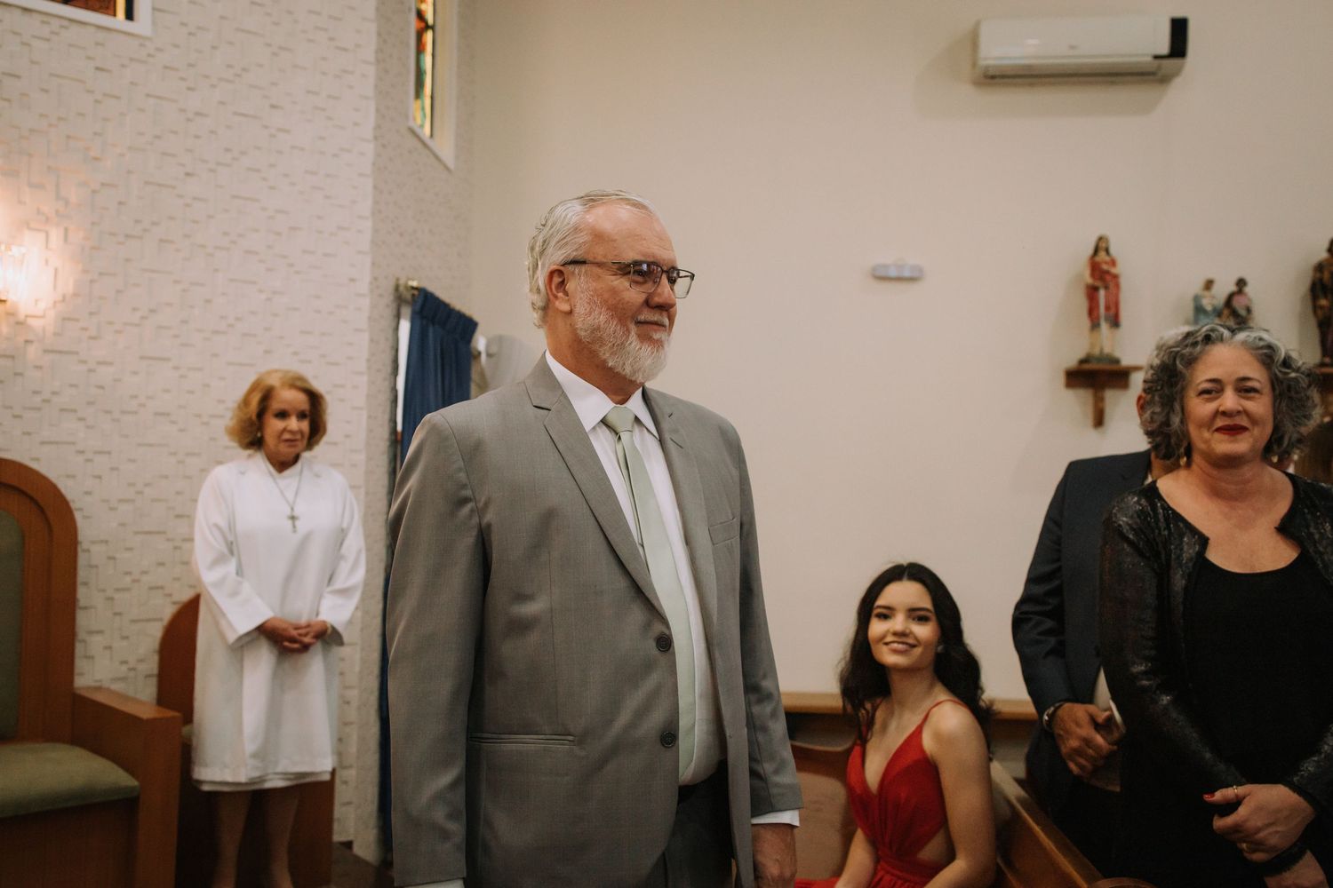 A candid moment at an indoor gathering with people standing and sitting in a room with textured walls and decor.