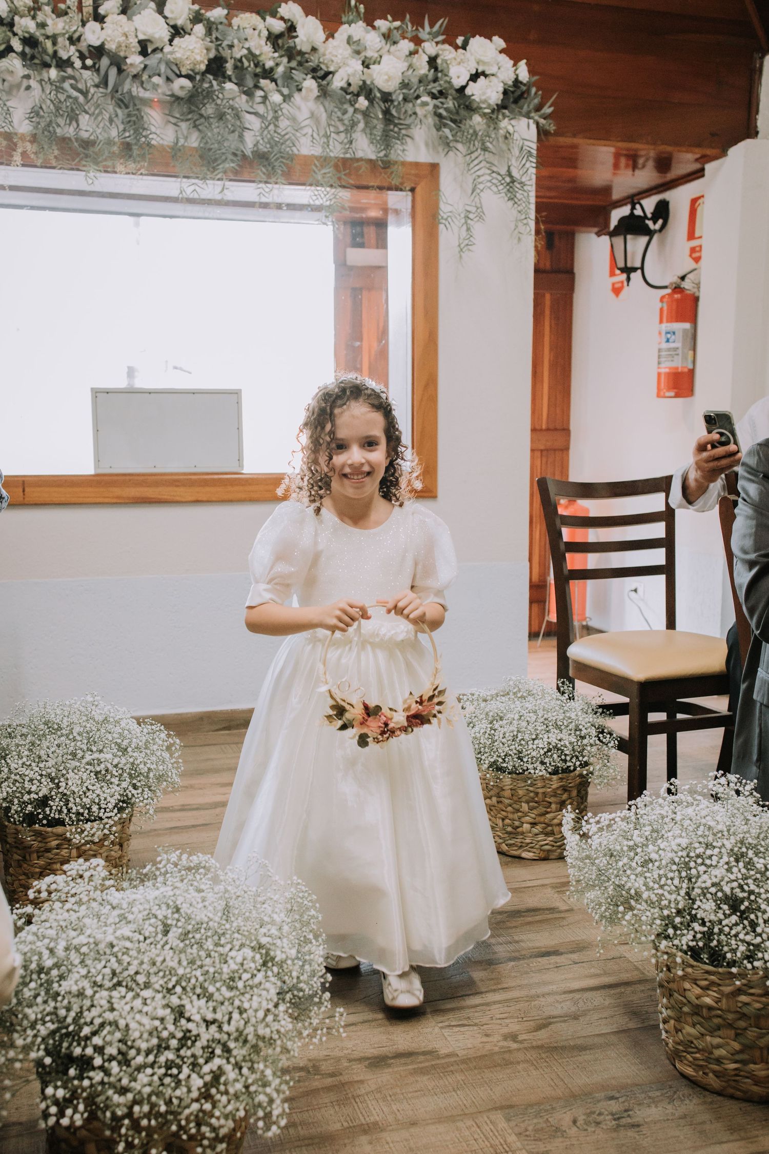 A young flower girl in a white dress plays near rustic baskets filled with baby's breath flowers on a wooden floor.