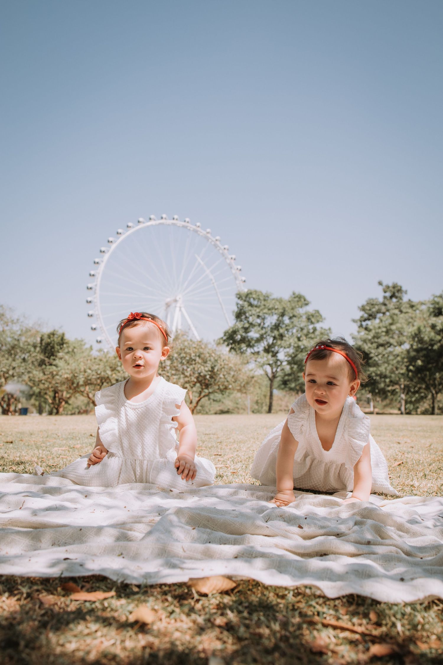 A baby in white dress crawls on a blanket outdoors with trees and blue sky in the background.