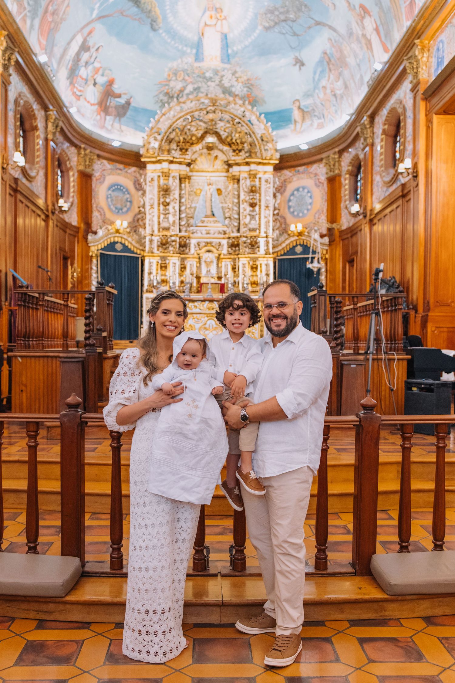 Family posing for a baptism photo inside an ornate church with a golden altar and painted ceiling.
