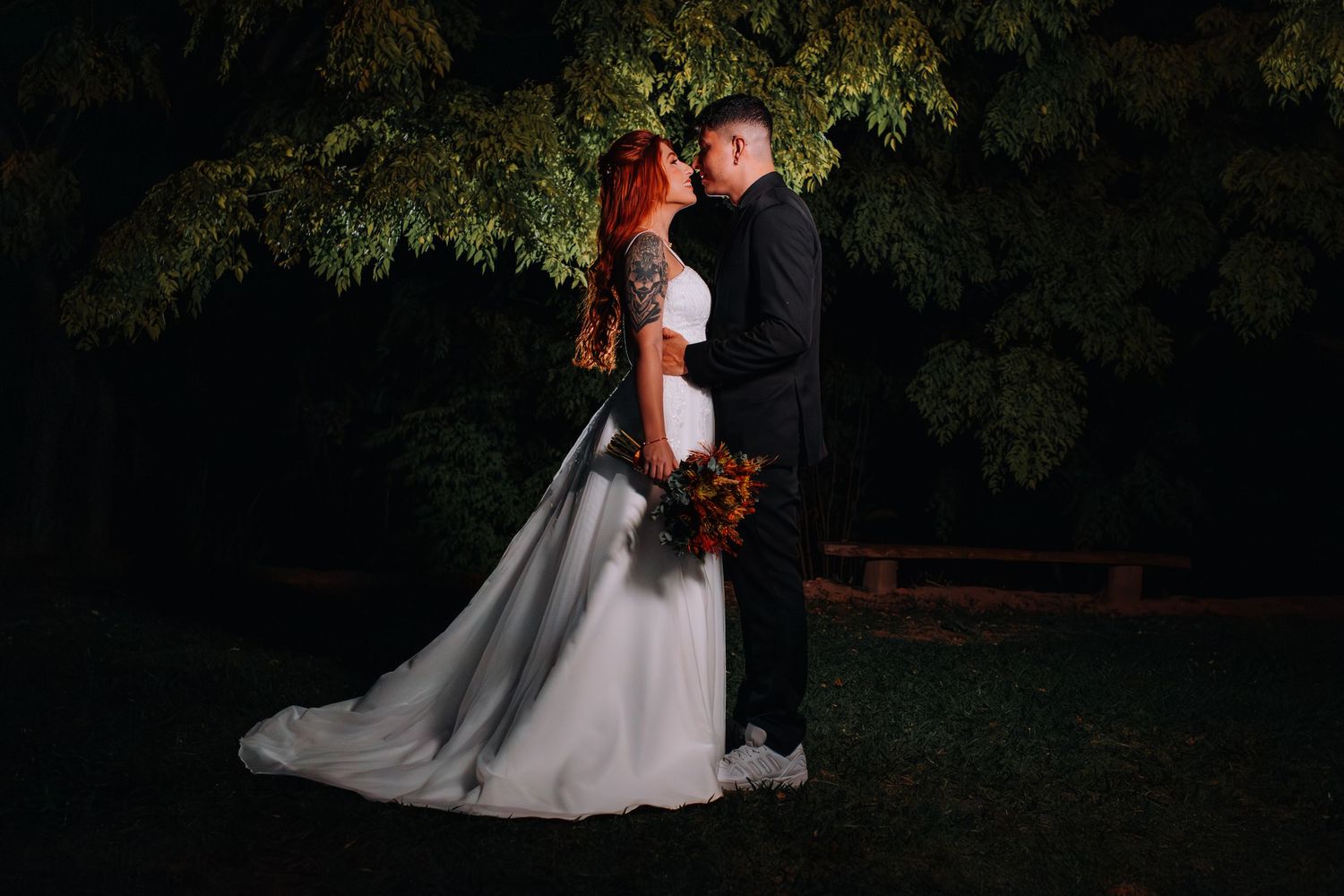 A romantic wedding couple embraces under a dark canopy of trees in an outdoor garden setting.