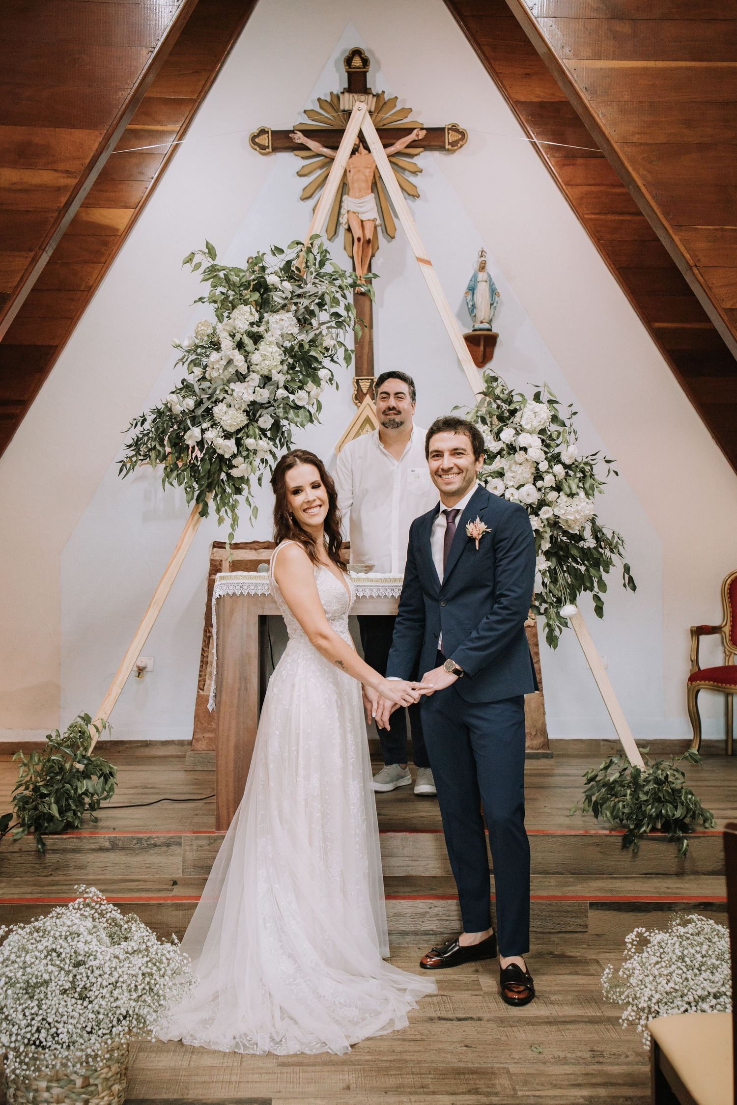 Elegant wedding couple poses in front of wooden cross and floral arrangements in rustic indoor venue.
