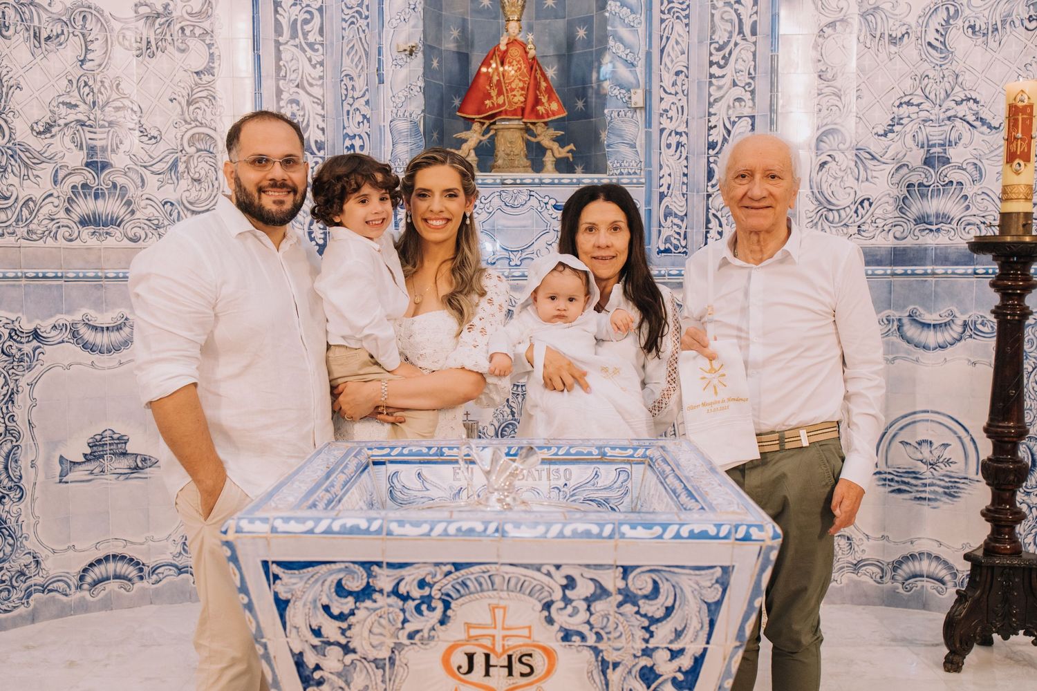 Family gathered around a baptismal font in a blue and white tiled church during a christening ceremony.