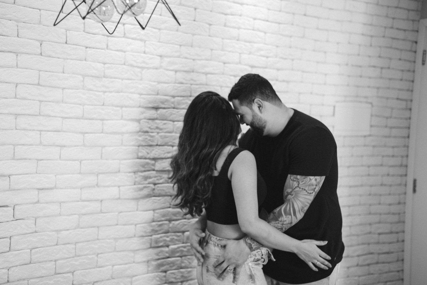 Romantic couple poses against white brick wall in casual attire.
