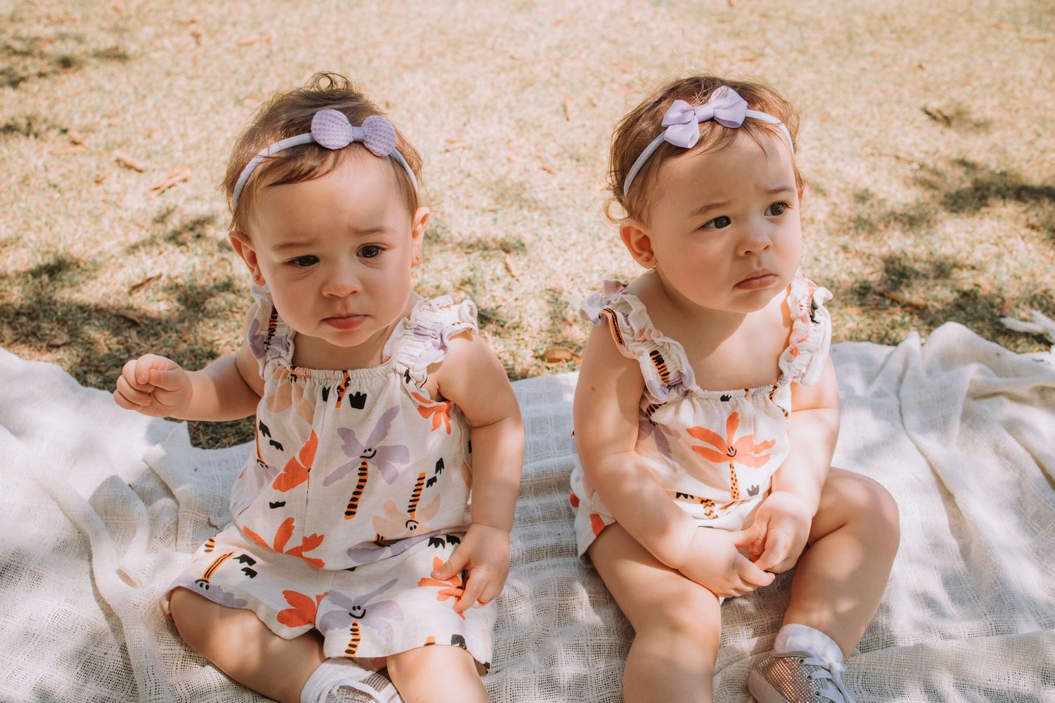 Two children in matching floral outfits sitting on a white blanket in a sunny field.
