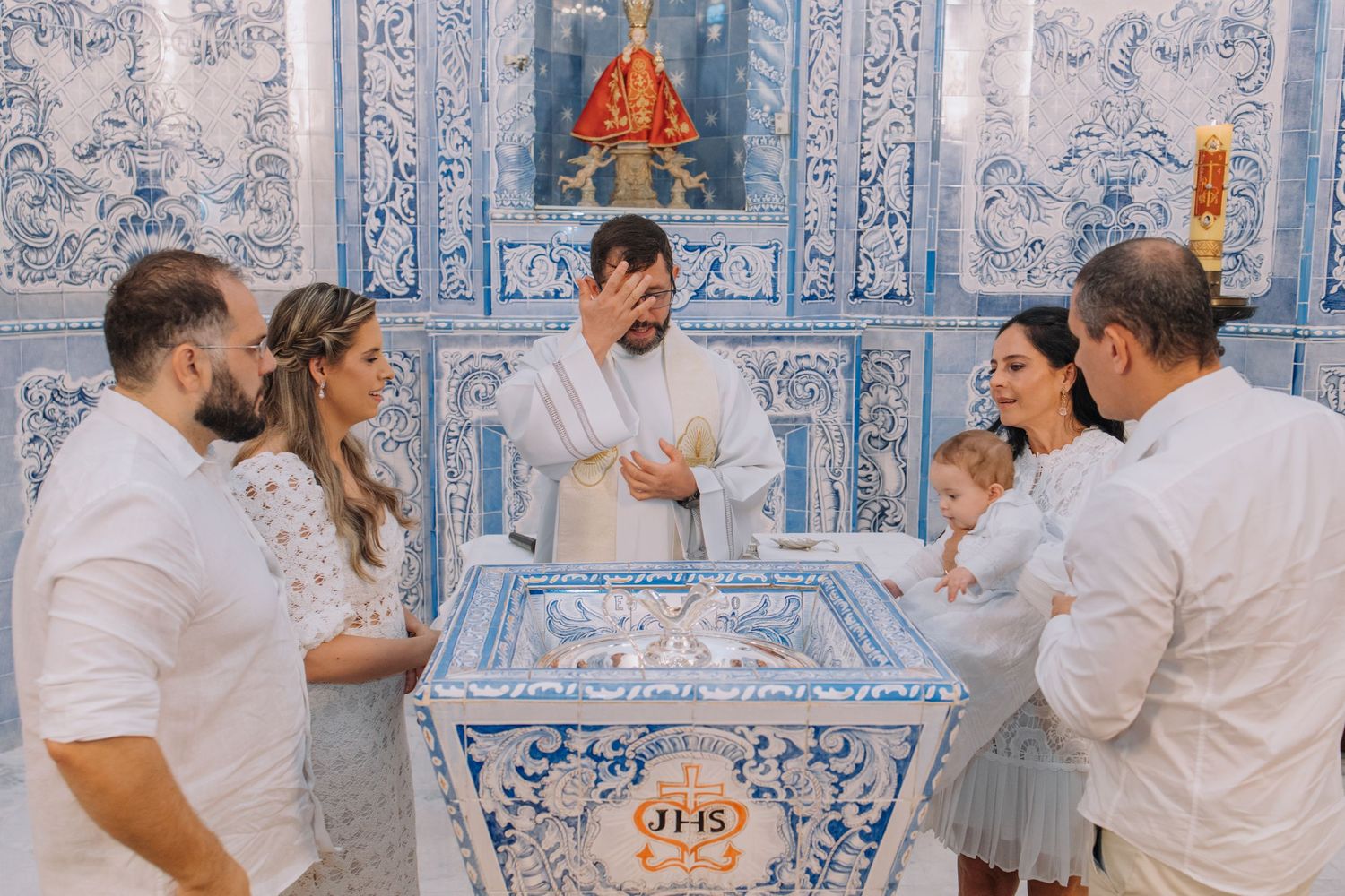 Catholic baptism ceremony with priest and family in white attire at ornate blue and white tiled chapel font.