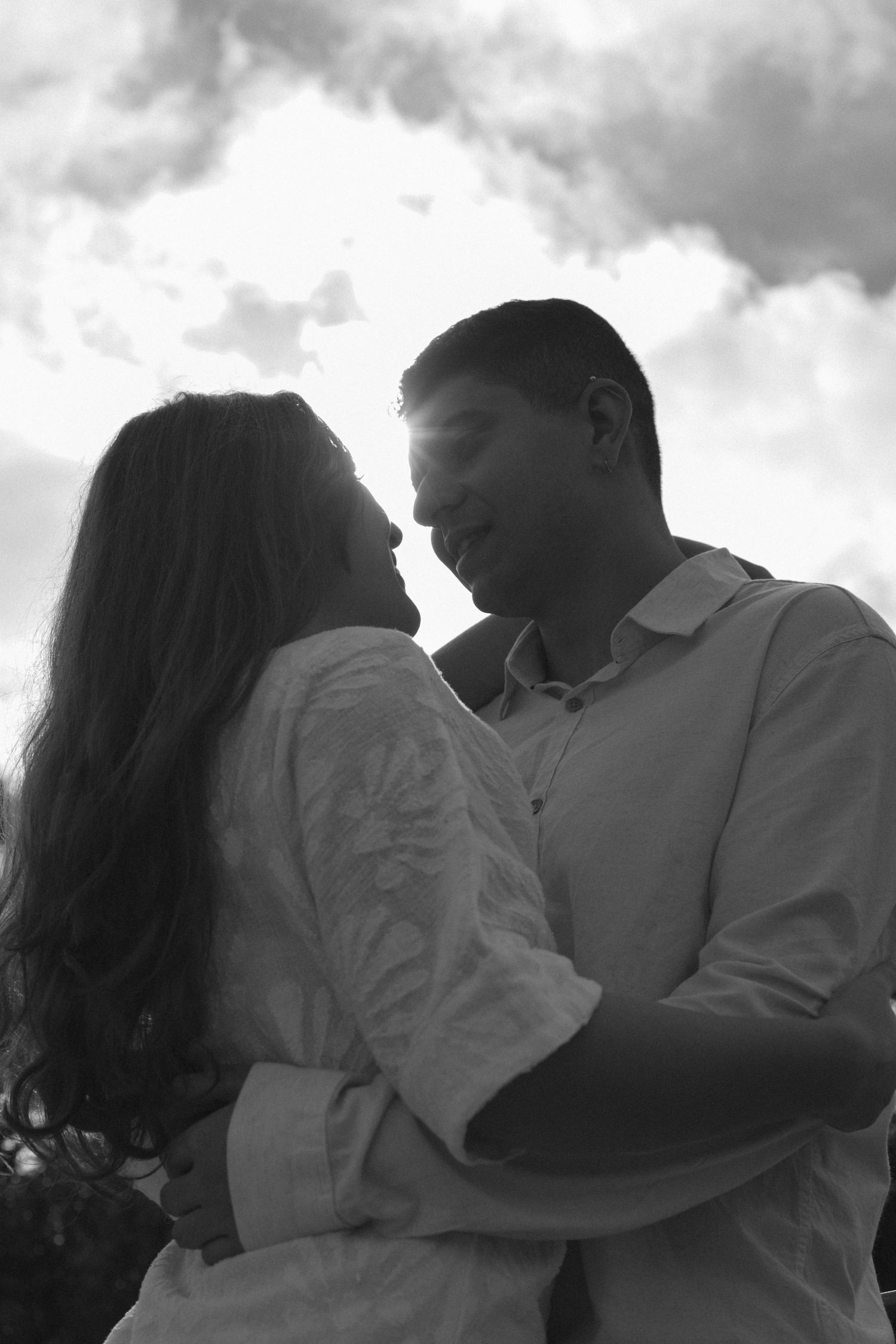 Black and white photo of a romantic couple embracing and about to kiss against a cloudy sky backdrop.