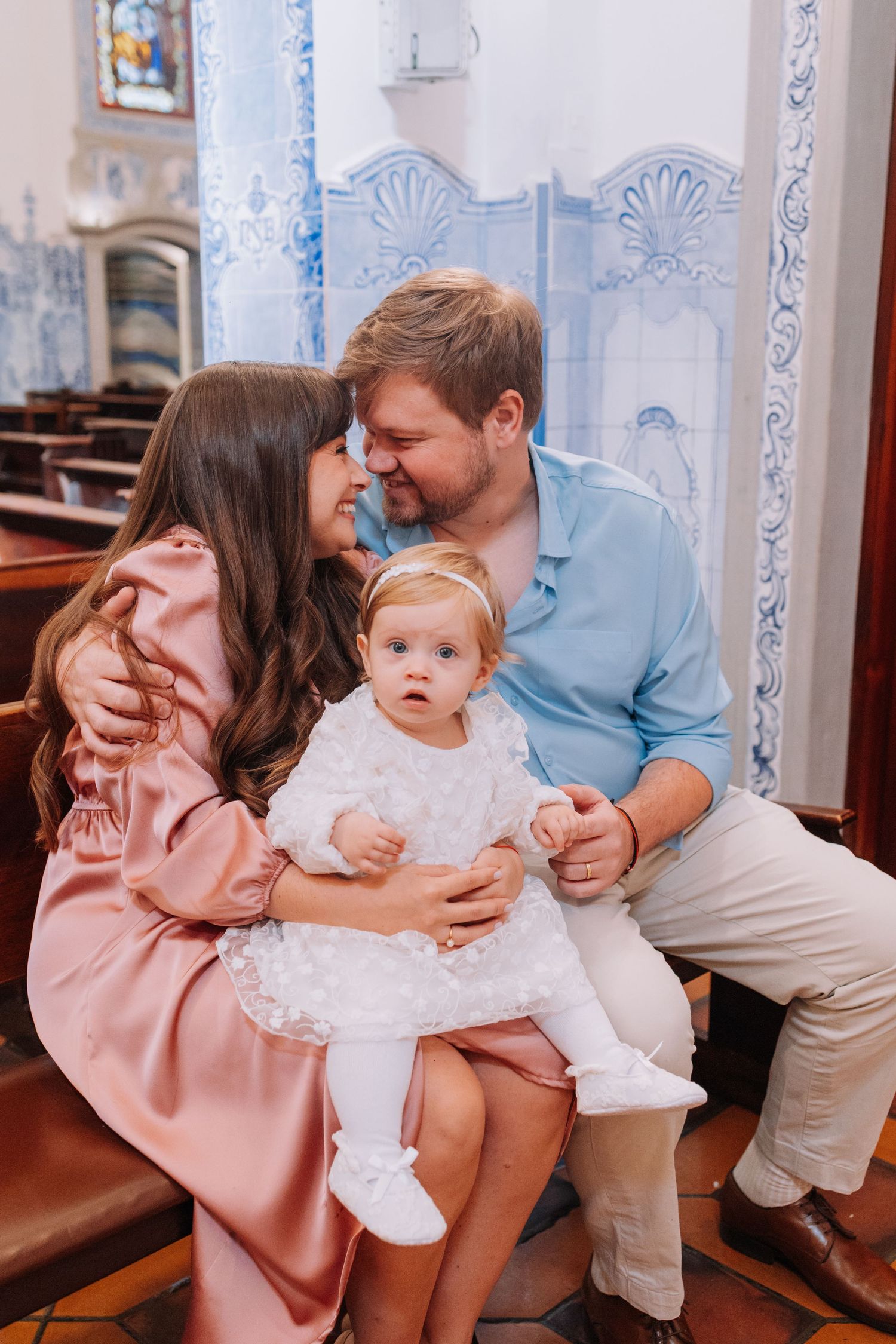 Family of three in a church, parents sharing a tender moment while holding their baby girl in a white dress.