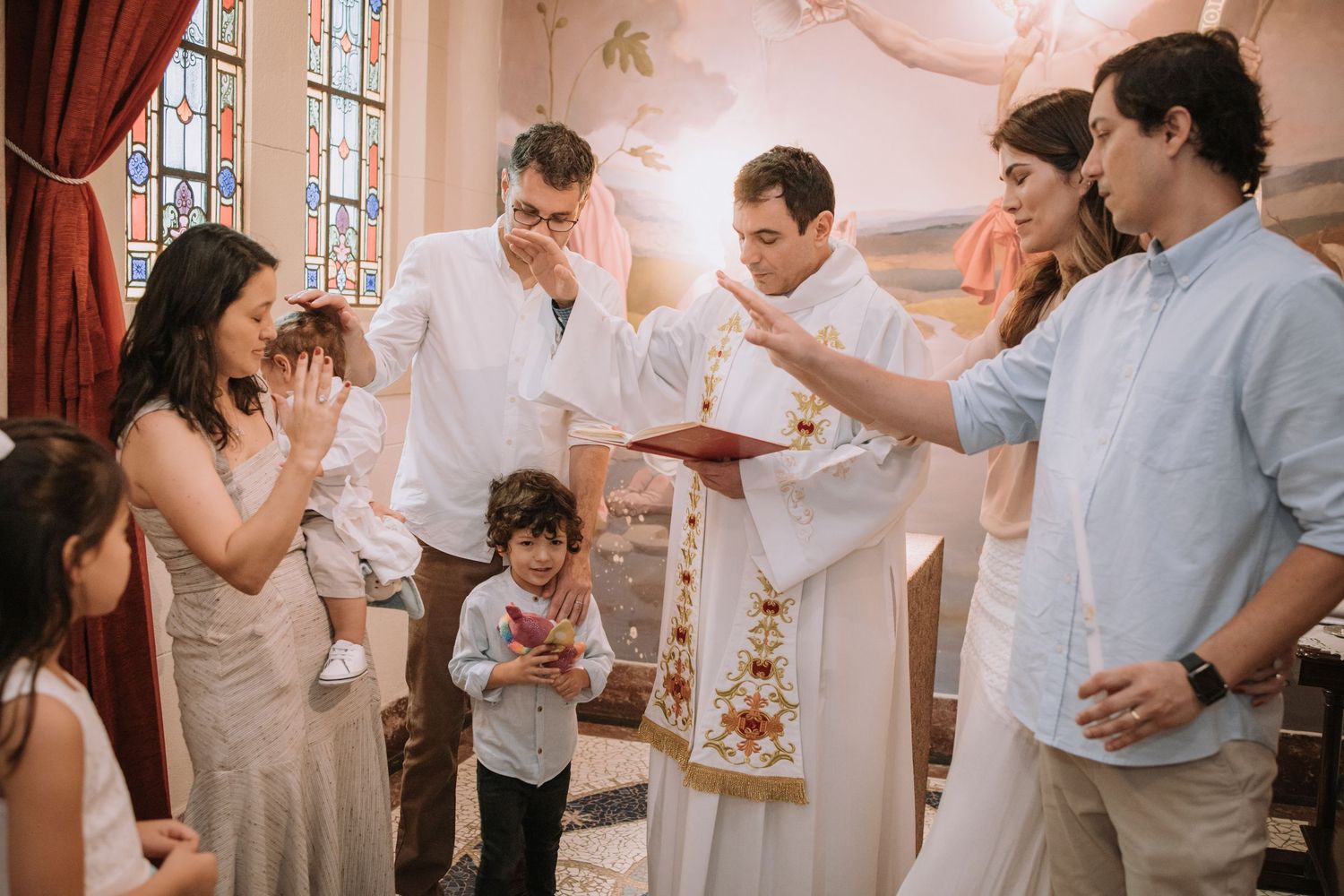 A religious ceremony takes place in a church with a priest and family gathered around a baptismal font.