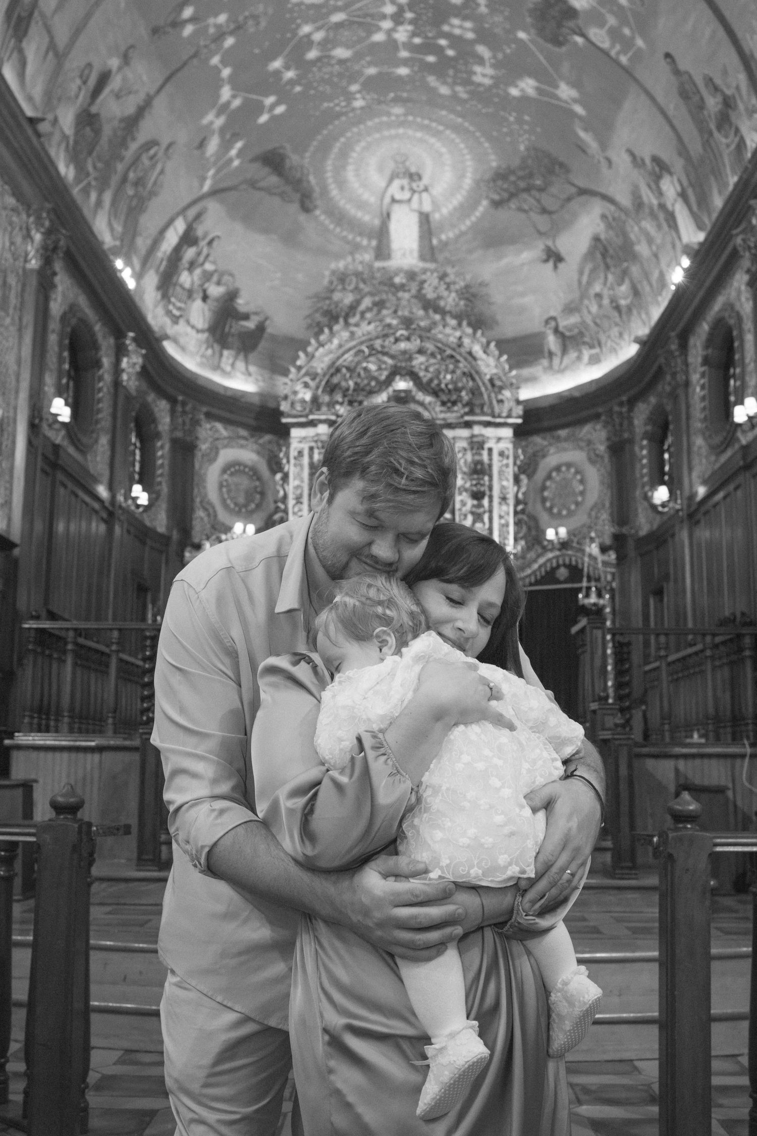 Black and white photo of parents holding baby in ornate church during christening ceremony.