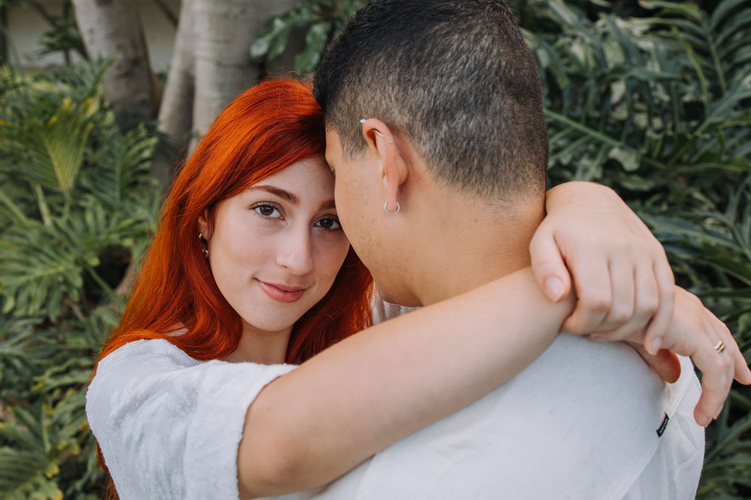 A redhead woman hugs a short-haired man outdoors, smiling softly amid lush green tropical plants.