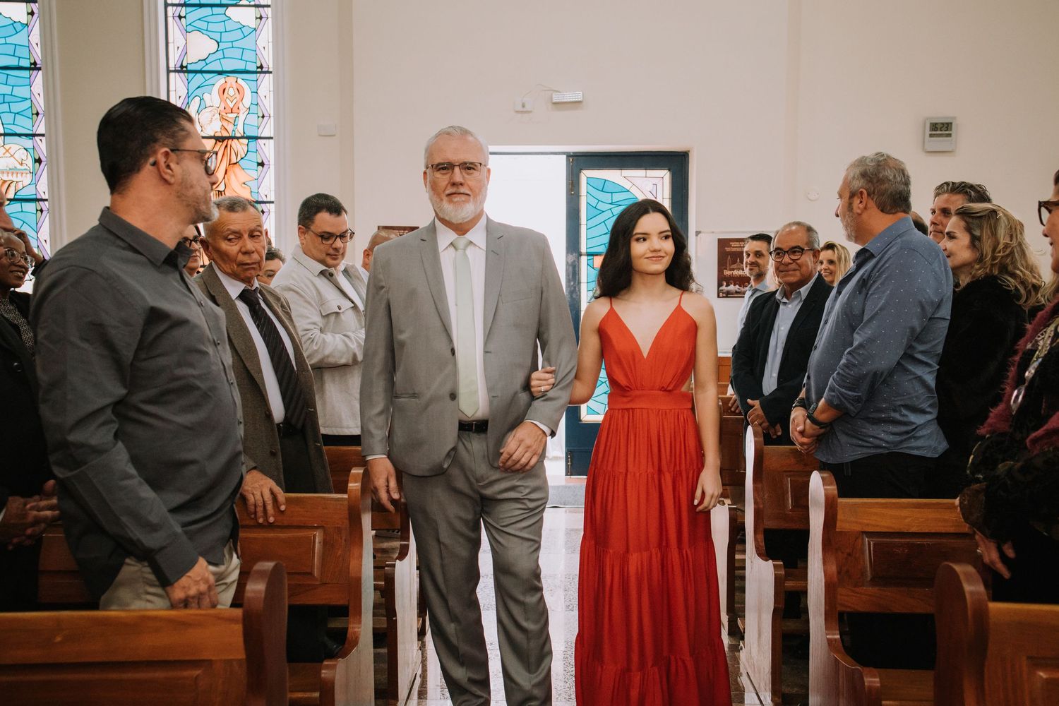 A group of wedding guests in formal attire gather inside a church with wooden pews and stained glass windows.