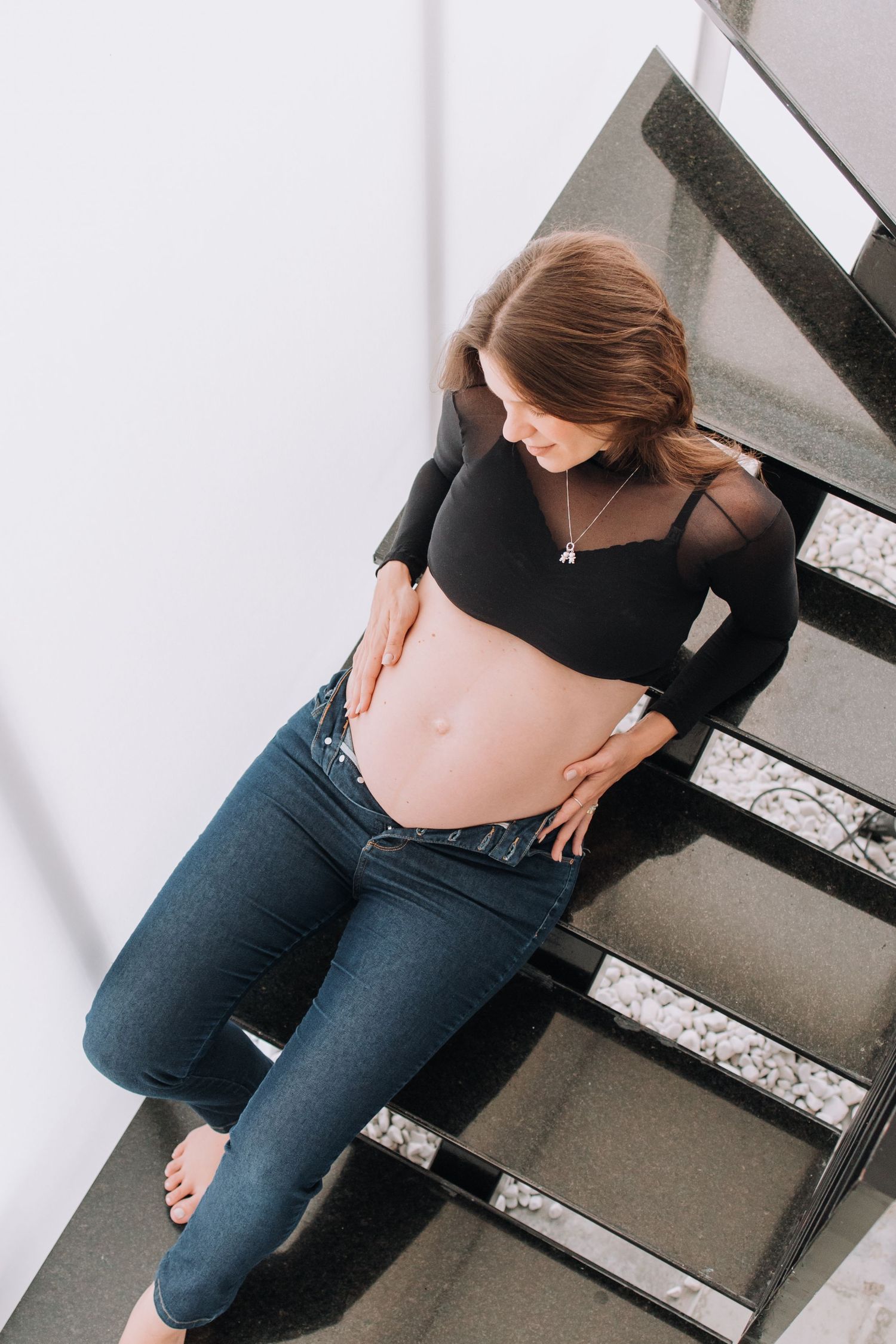 Pregnant woman in black top and jeans poses on modern staircase while holding belly.