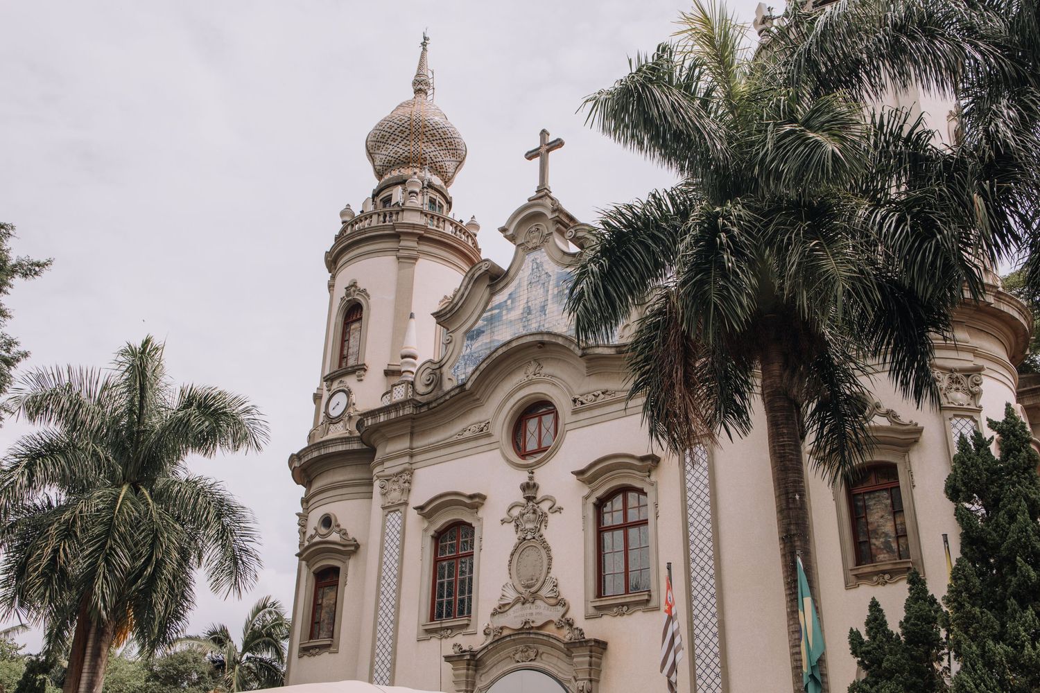 Baroque colonial church with ornate facade, cross-topped dome, and lush palm trees under overcast sky.