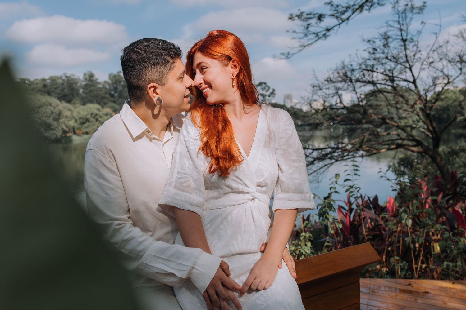 Couple in white outfits sharing a romantic moment outdoors near a lake, surrounded by lush greenery.
