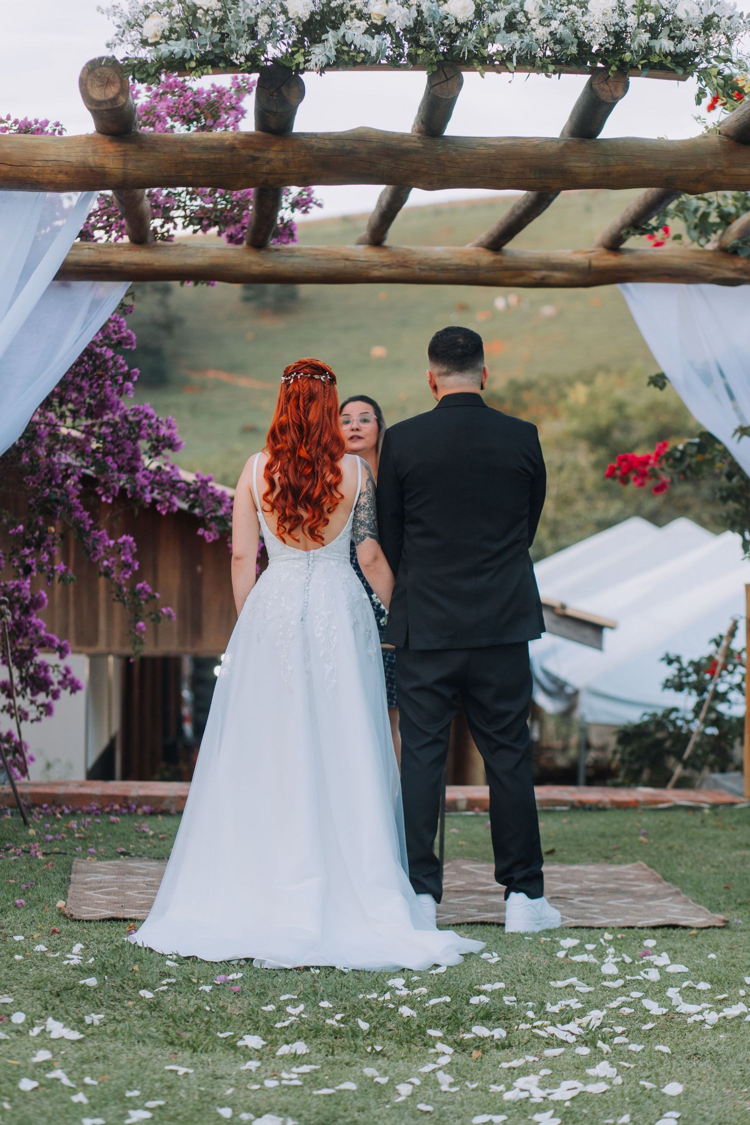 A couple stands at a rustic outdoor wedding altar adorned with purple flowers and white petals on the ground.