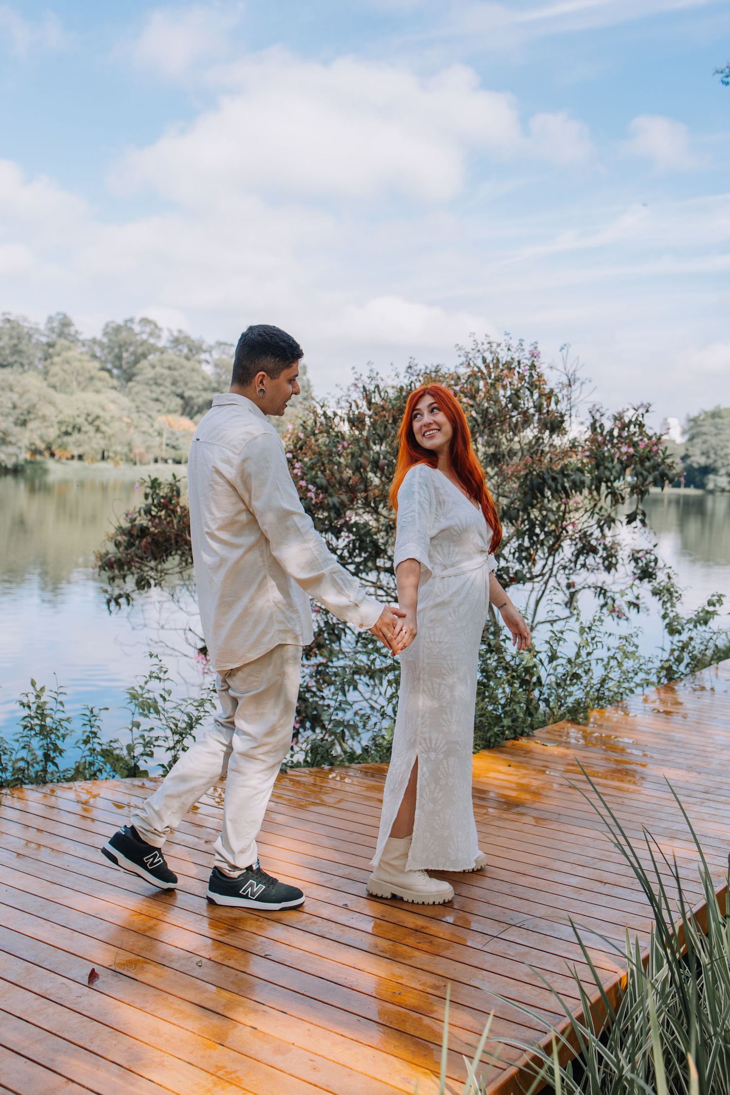 Couple in white outfits holding hands on a wooden dock by a serene lake surrounded by lush greenery.