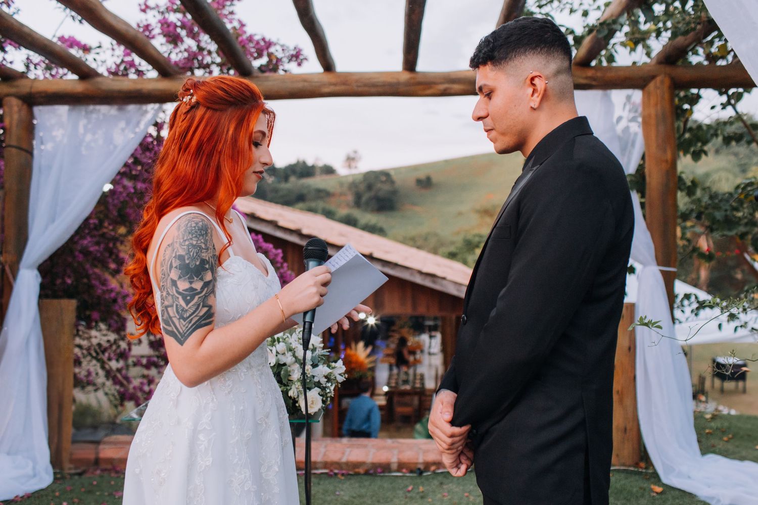 A couple exchanges vows during an outdoor wedding ceremony under a wooden arch decorated with flowers and white fabric.