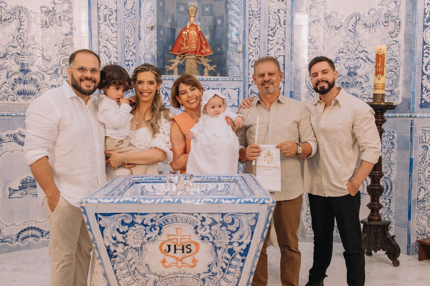 Family gathered around a baptismal font in a church adorned with blue and white tiles during a christening ceremony.