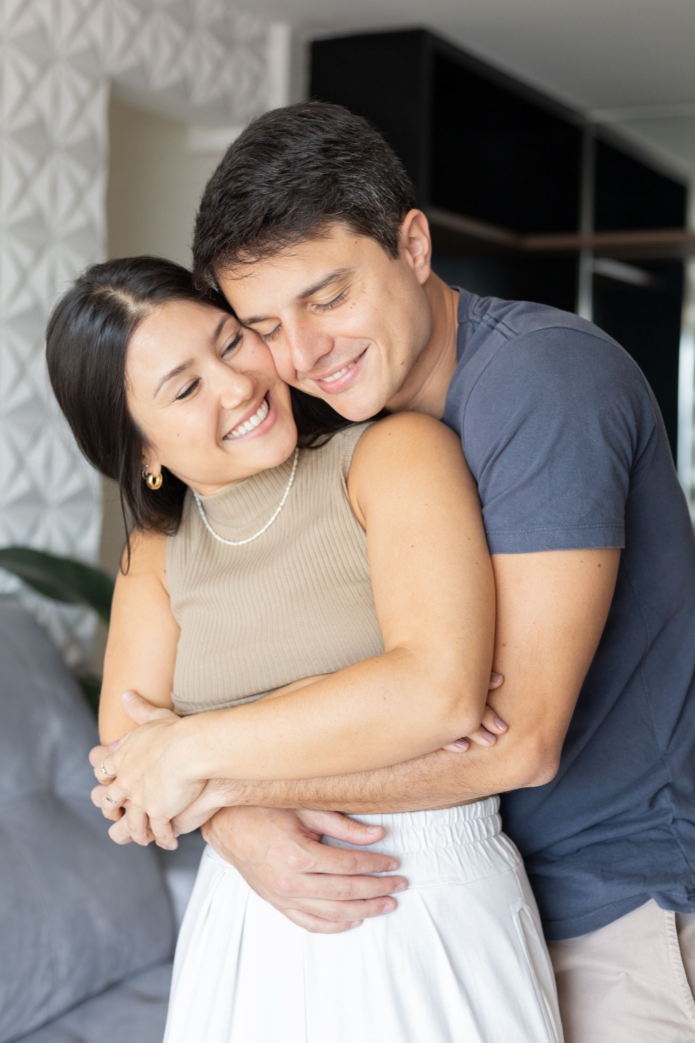 Happy couple embracing at home, smiling woman in beige top held by man in gray t-shirt in cozy living room.