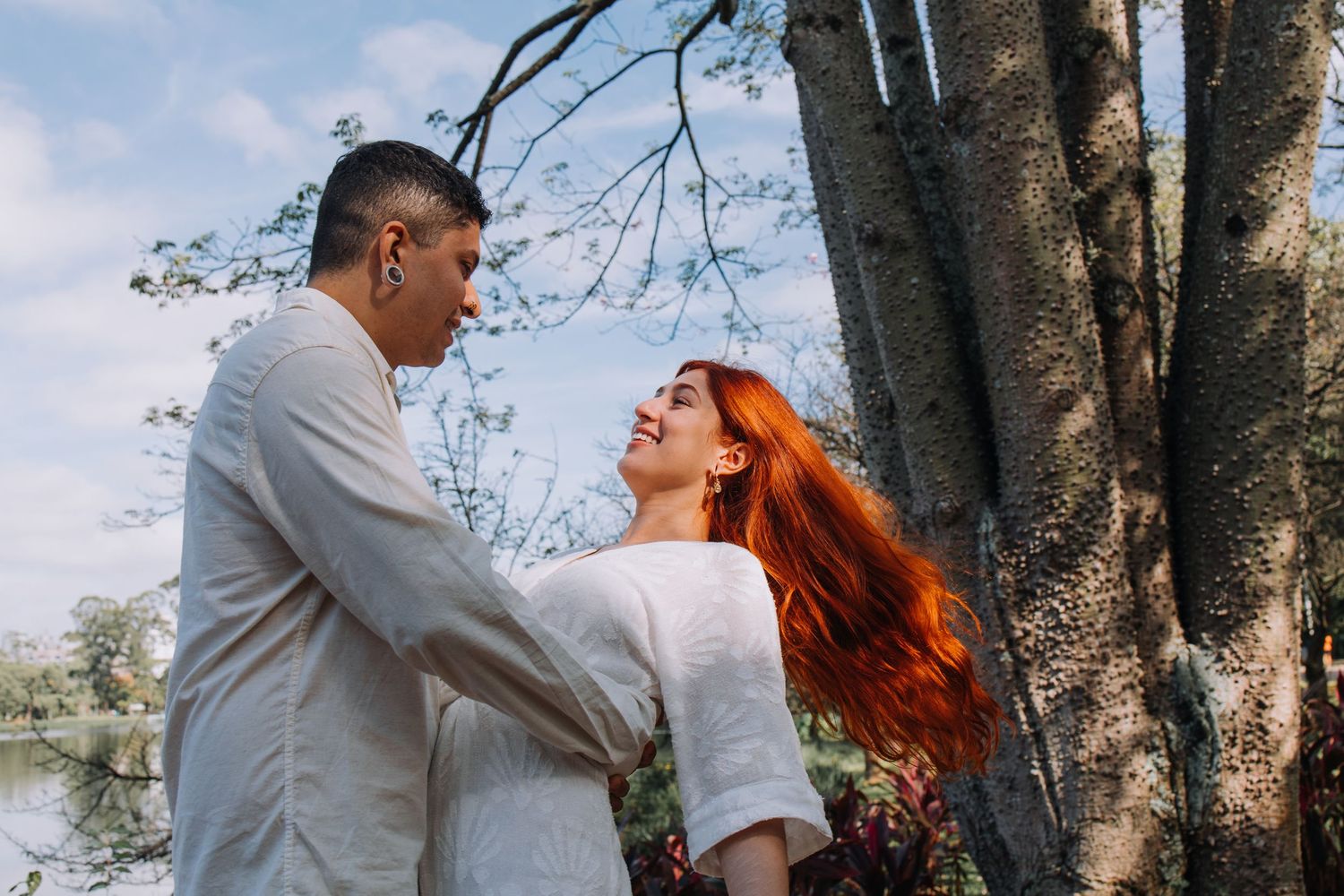 A couple holds hands and gazes at each other near trees, the woman with long red hair smiling up at her partner.