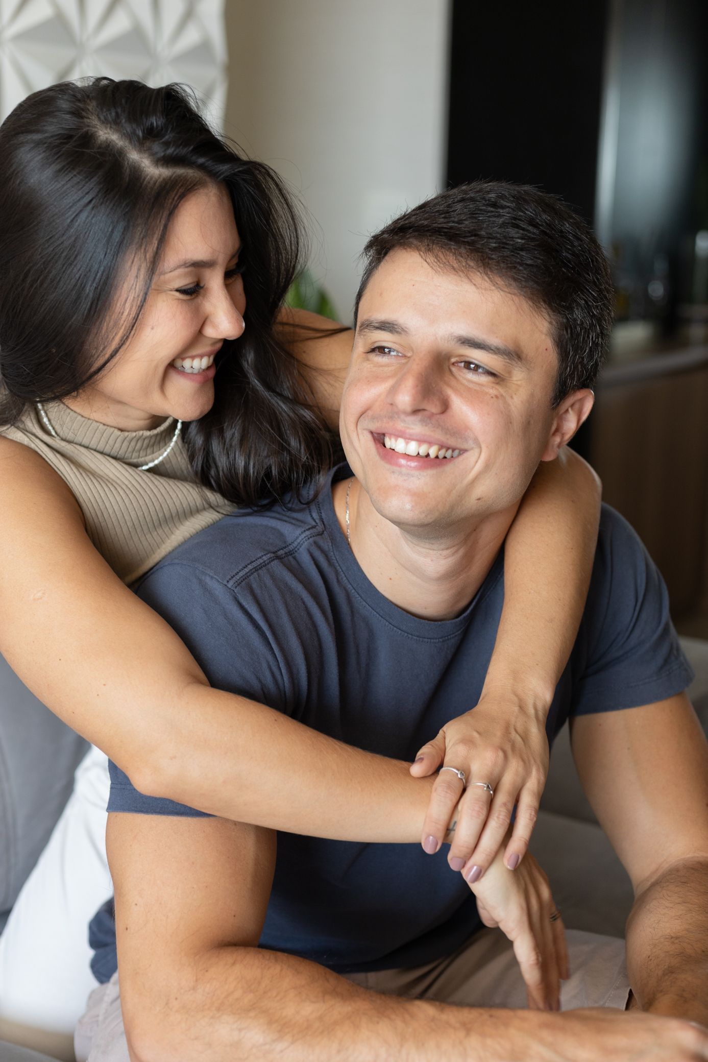 Happy couple smiling and embracing at home, woman hugging man from behind in a cozy indoor setting.