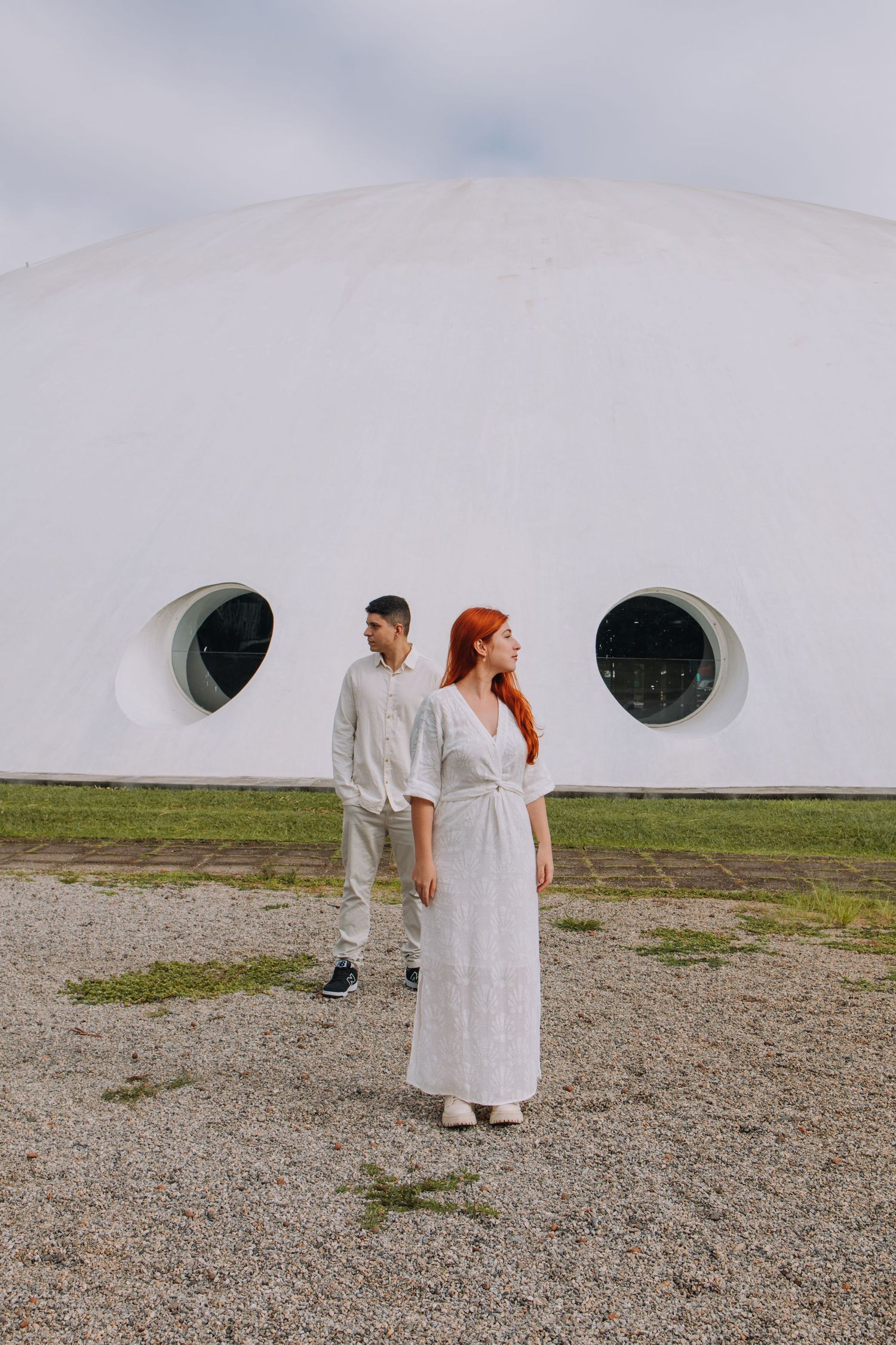 A couple in white outfits stands before a modern white dome building with circular windows on a cloudy day.