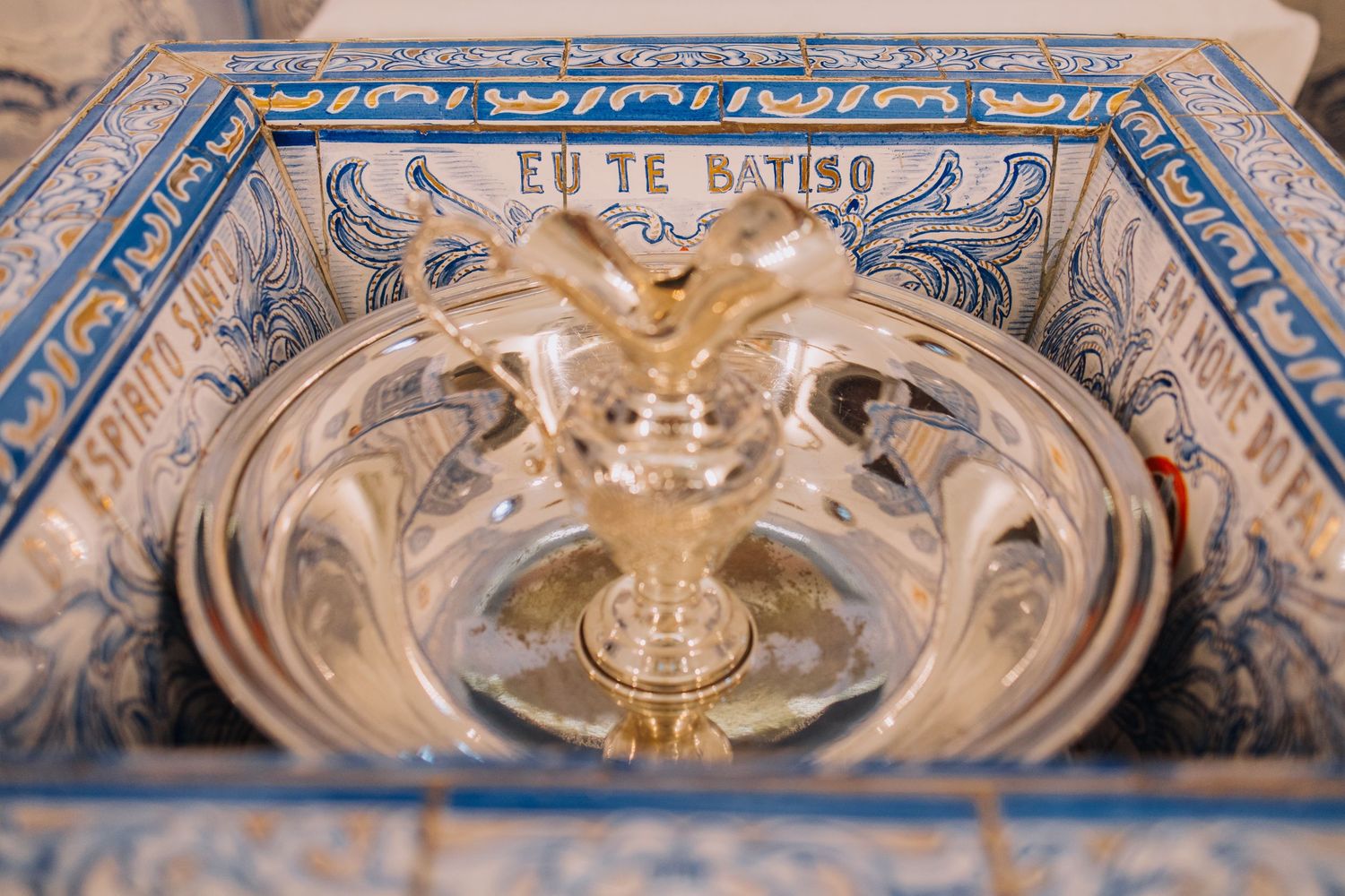 Ornate baptismal font with glass bowl, gold accents, and blue Portuguese azulejo tiles inscribed EU TE BATISO.