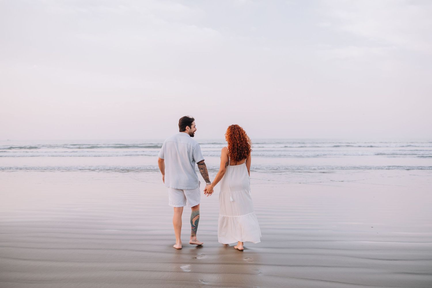 Couple walking hand in hand along a misty beach shoreline at sunset.