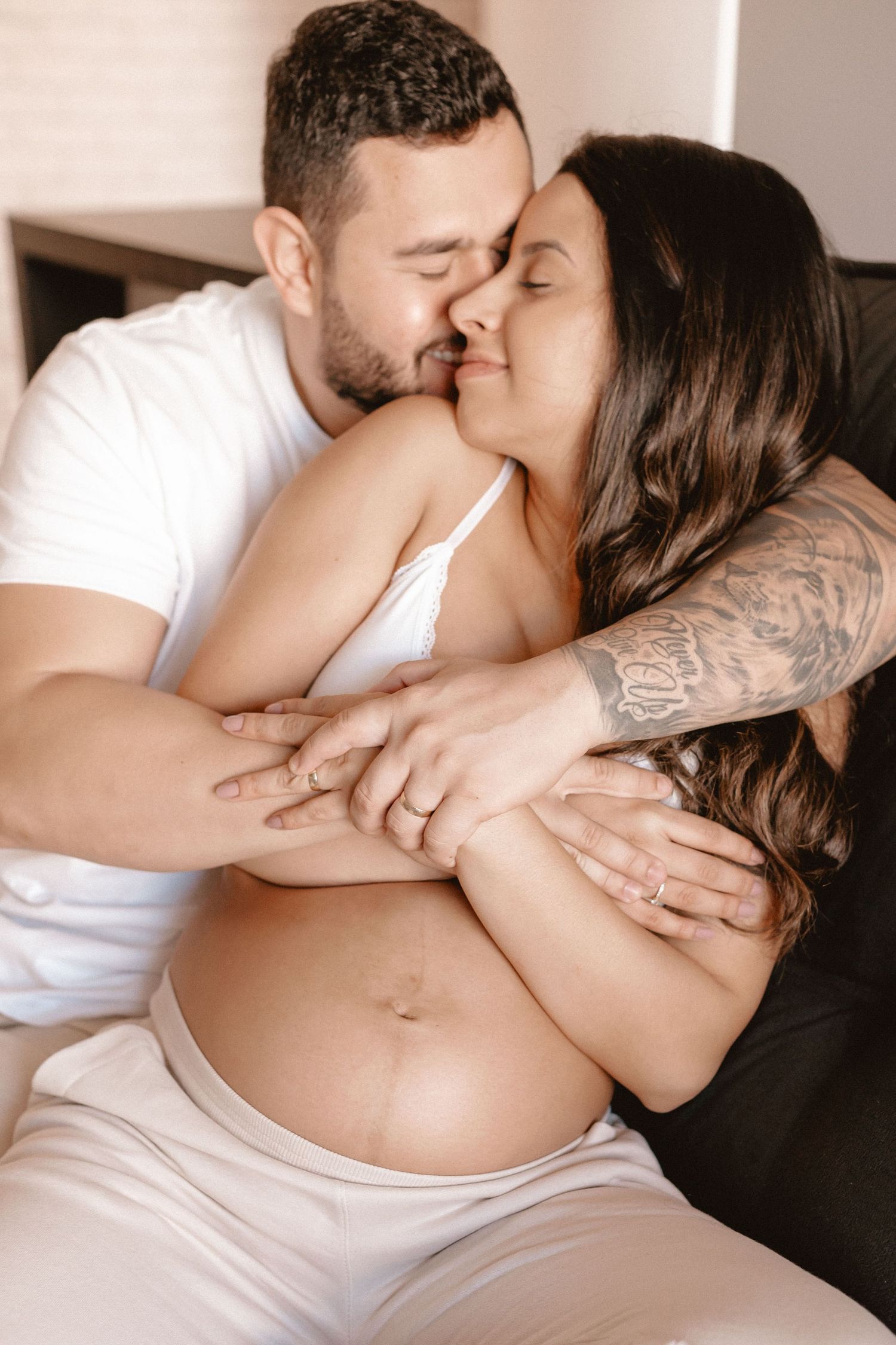 Intimate maternity photoshoot with couple in white clothing on light background.