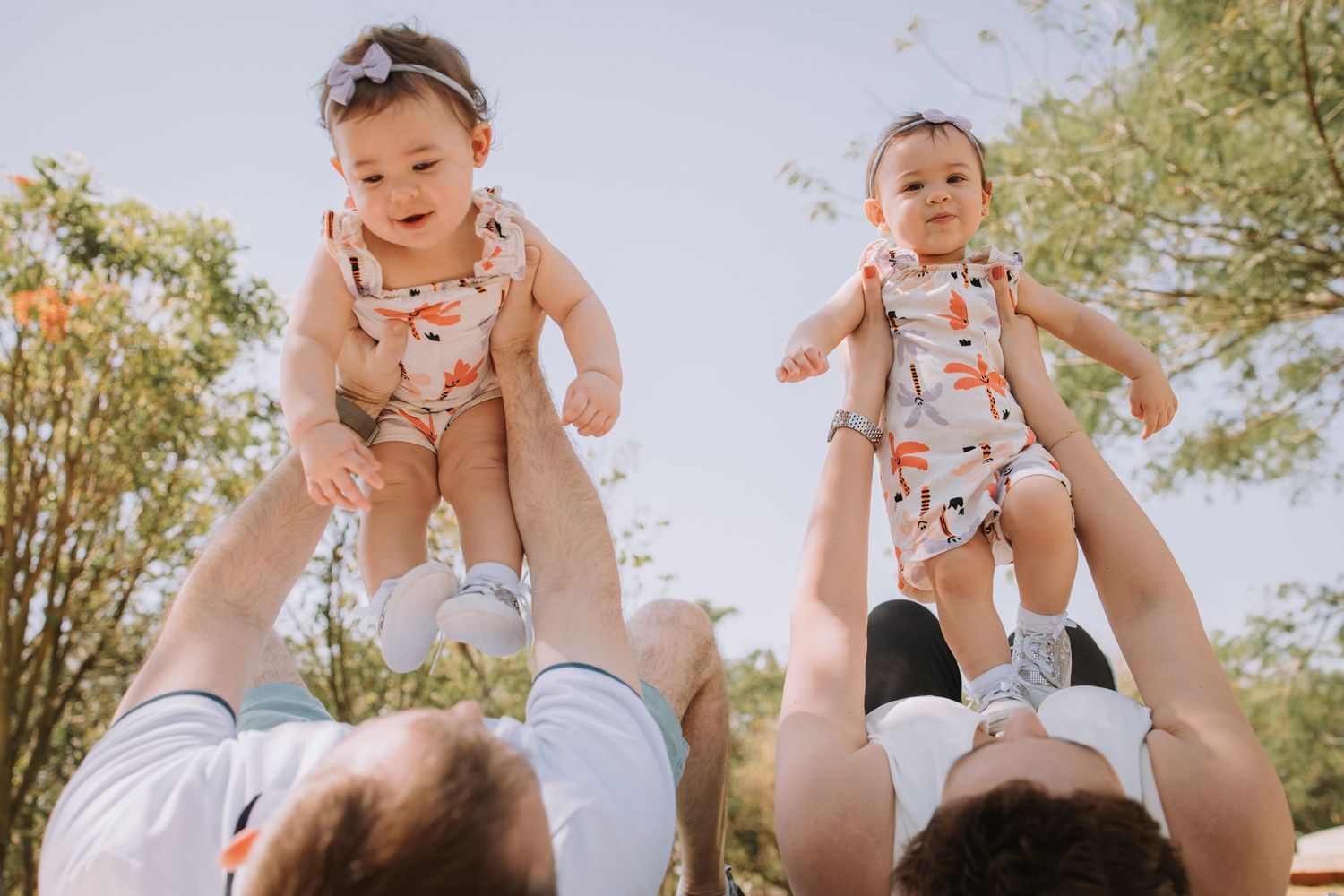 Parents lifting and kissing baby outdoors in sunny garden setting.