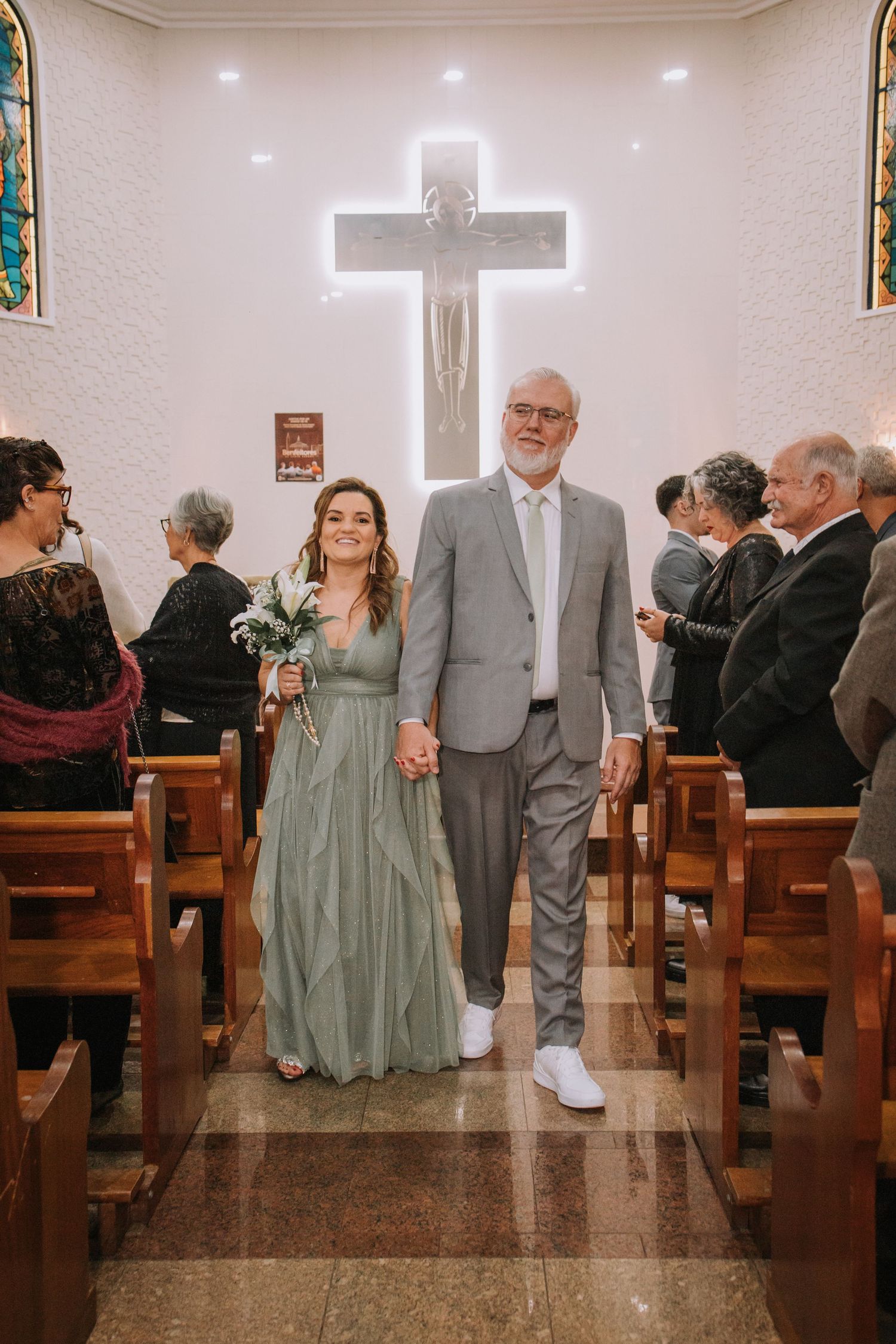 Wedding couple in sage green dress and gray suit pose together in church with illuminated cross in background.
