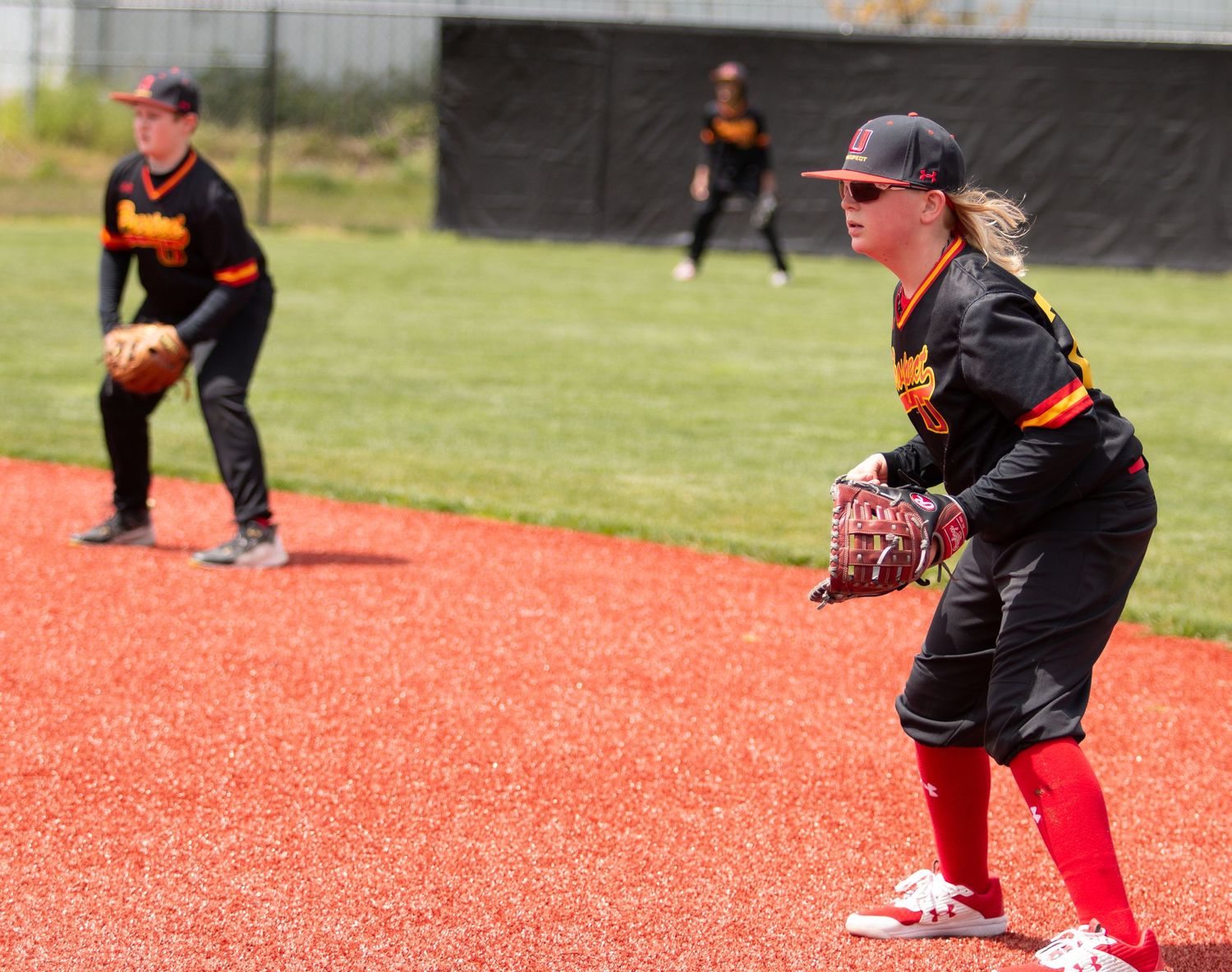 Baseball players in dark uniforms practicing defensive positions on red infield dirt.