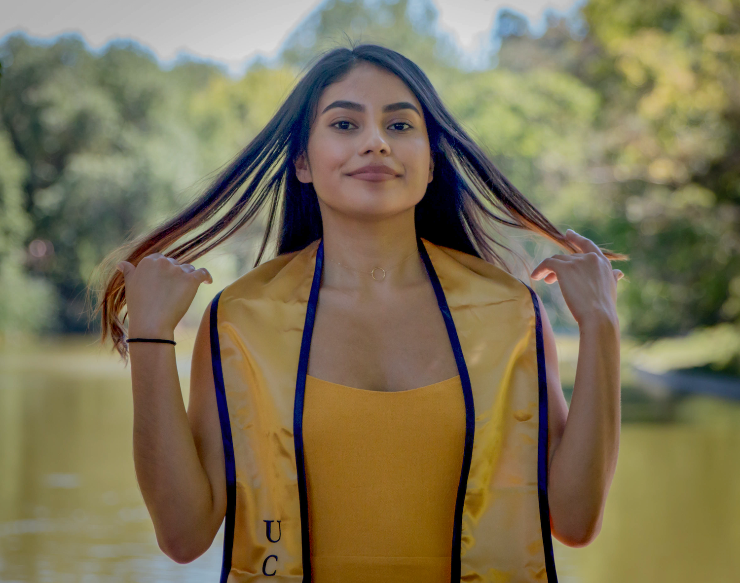 Graduate in yellow dress poses with graduation stole in outdoor park setting.