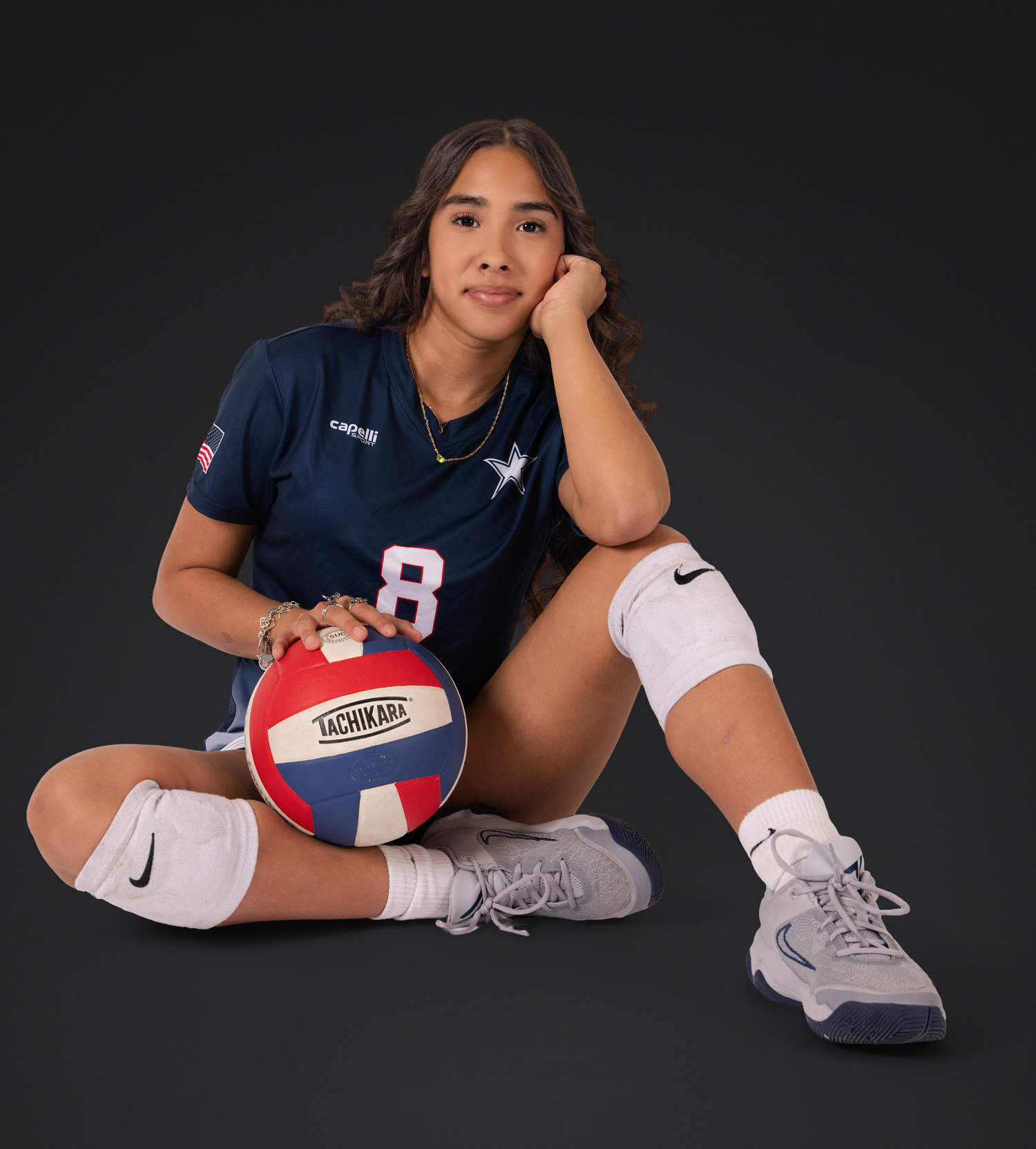Athlete in navy jersey poses with volleyball and knee pads on dark background.