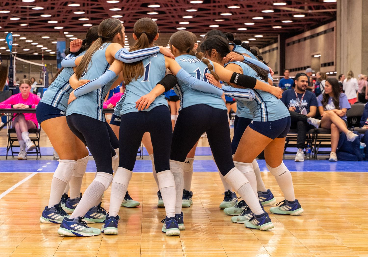 Indoor volleyball players in light blue uniforms positioned defensively on a wooden court.