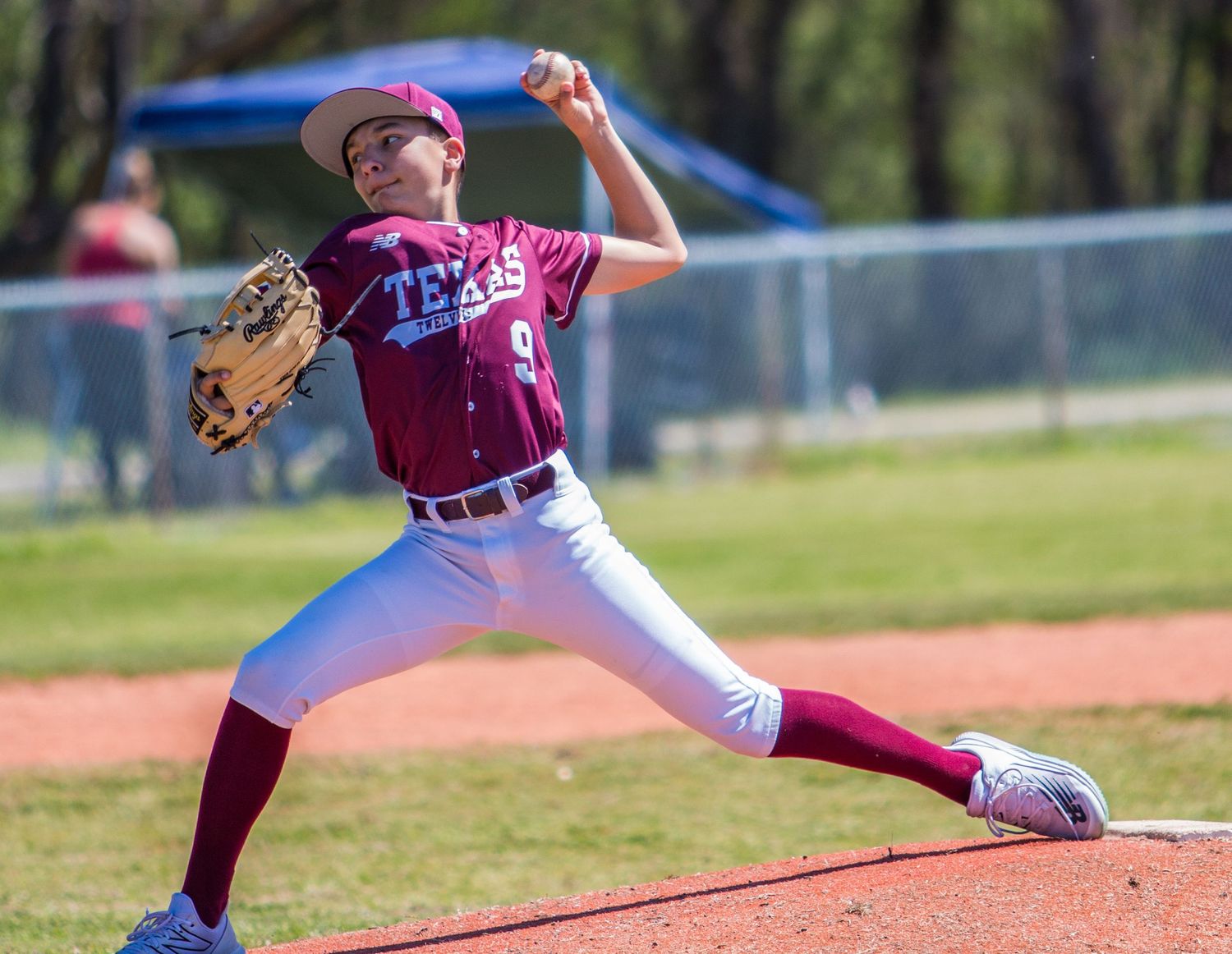Youth baseball pitcher in maroon uniform winds up to throw pitch during game.