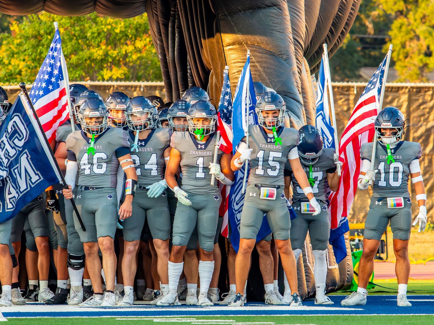 Football team members stand in a line during pre-game ceremony with American flags waving in the background.