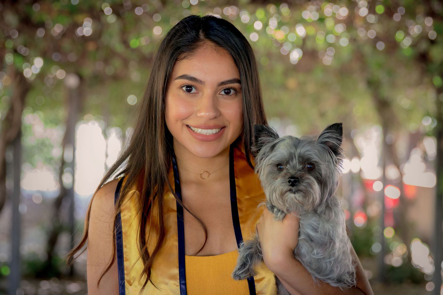 Graduate poses with small grey dog during outdoor graduation photoshoot with bokeh effect.