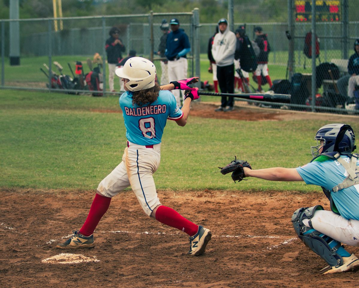 Youth baseball player in white and red uniform swings bat at home plate while catcher crouches in teal uniform.