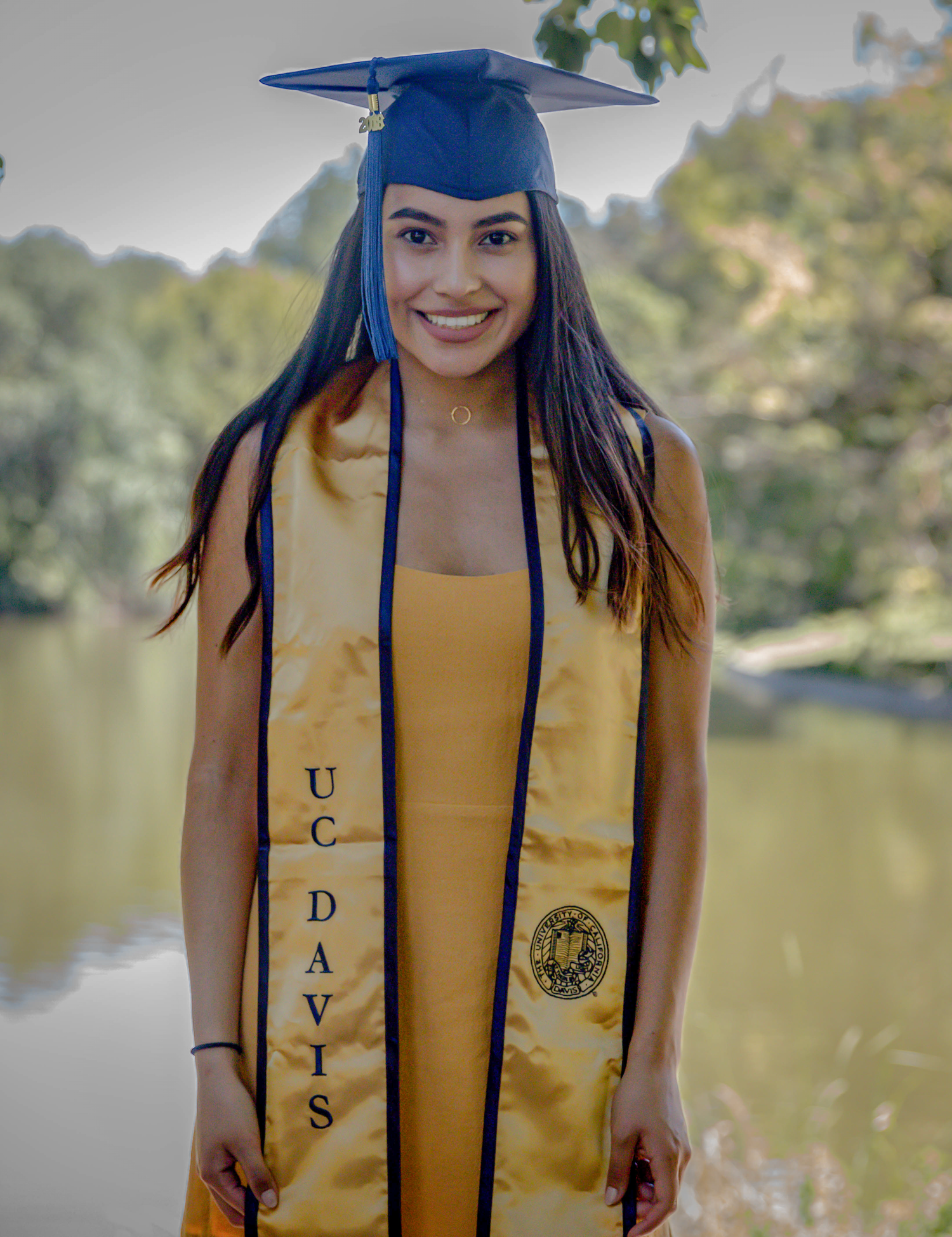 Graduate in yellow dress with blue cap and stole poses by lake during graduation photos.