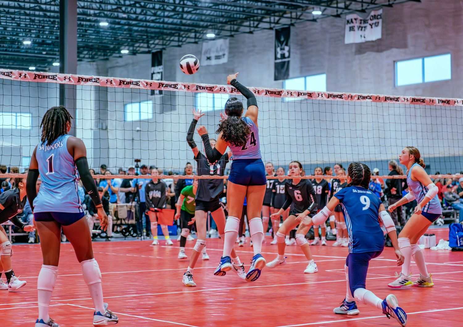 Volleyball players compete at net during tournament on red court.