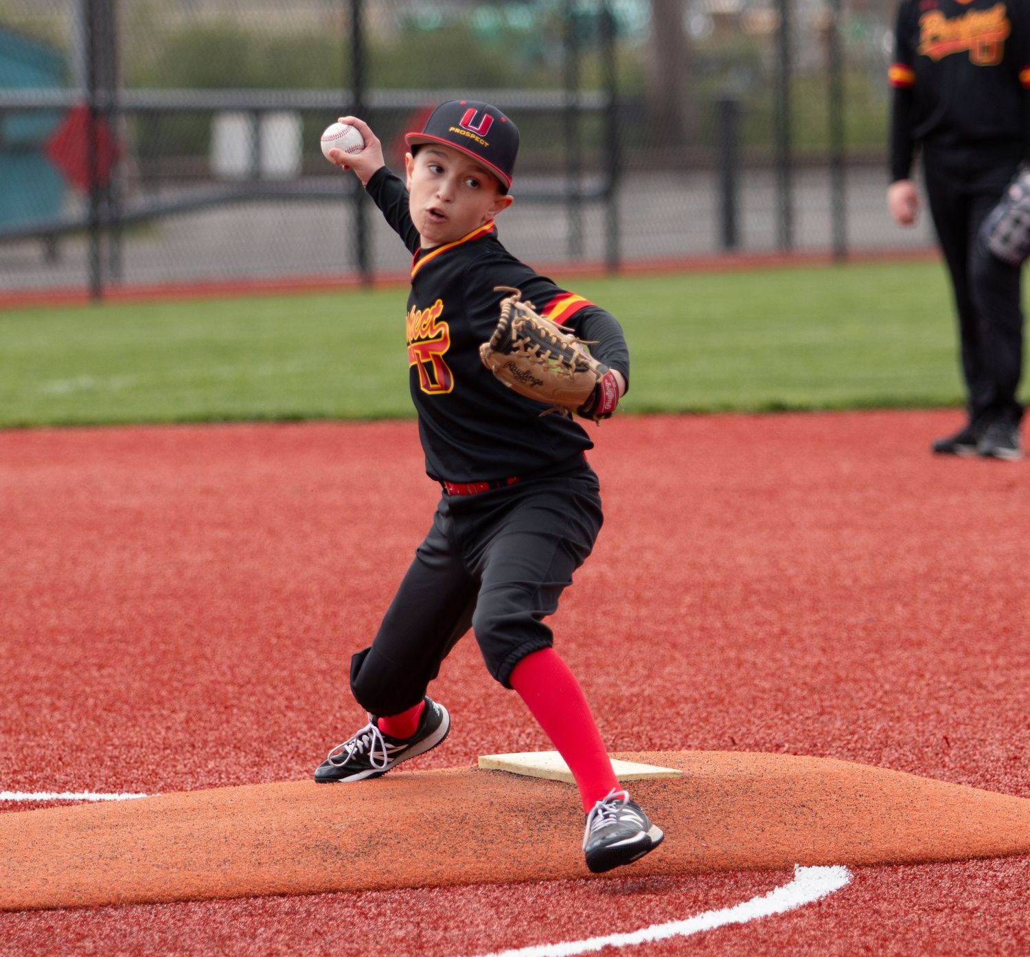Youth baseball pitcher in black uniform and red socks delivers pitch from mound on red turf field.