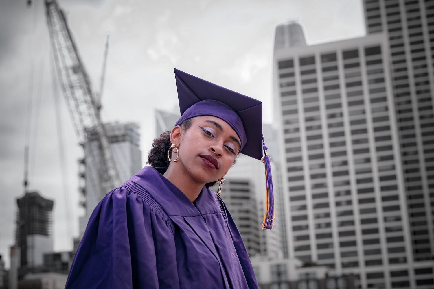 Graduate in purple regalia poses against urban San Francisco skyline backdrop.