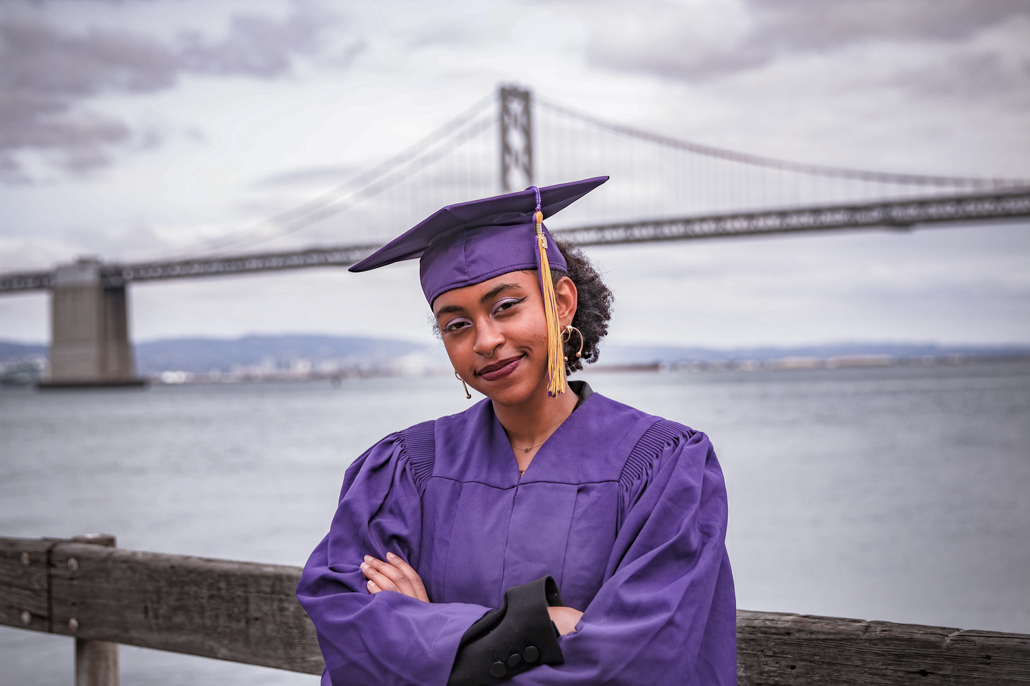 Graduate in purple cap and gown poses with San Francisco Bay Bridge in background.