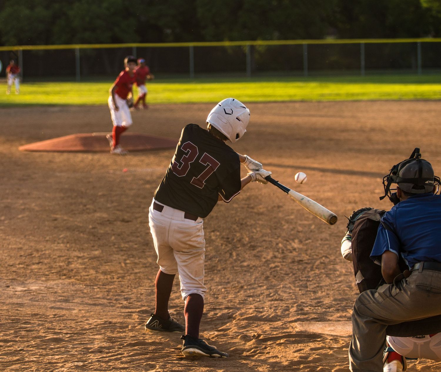 Baseball batter swings at pitch during sunset game with catcher and umpire in frame.